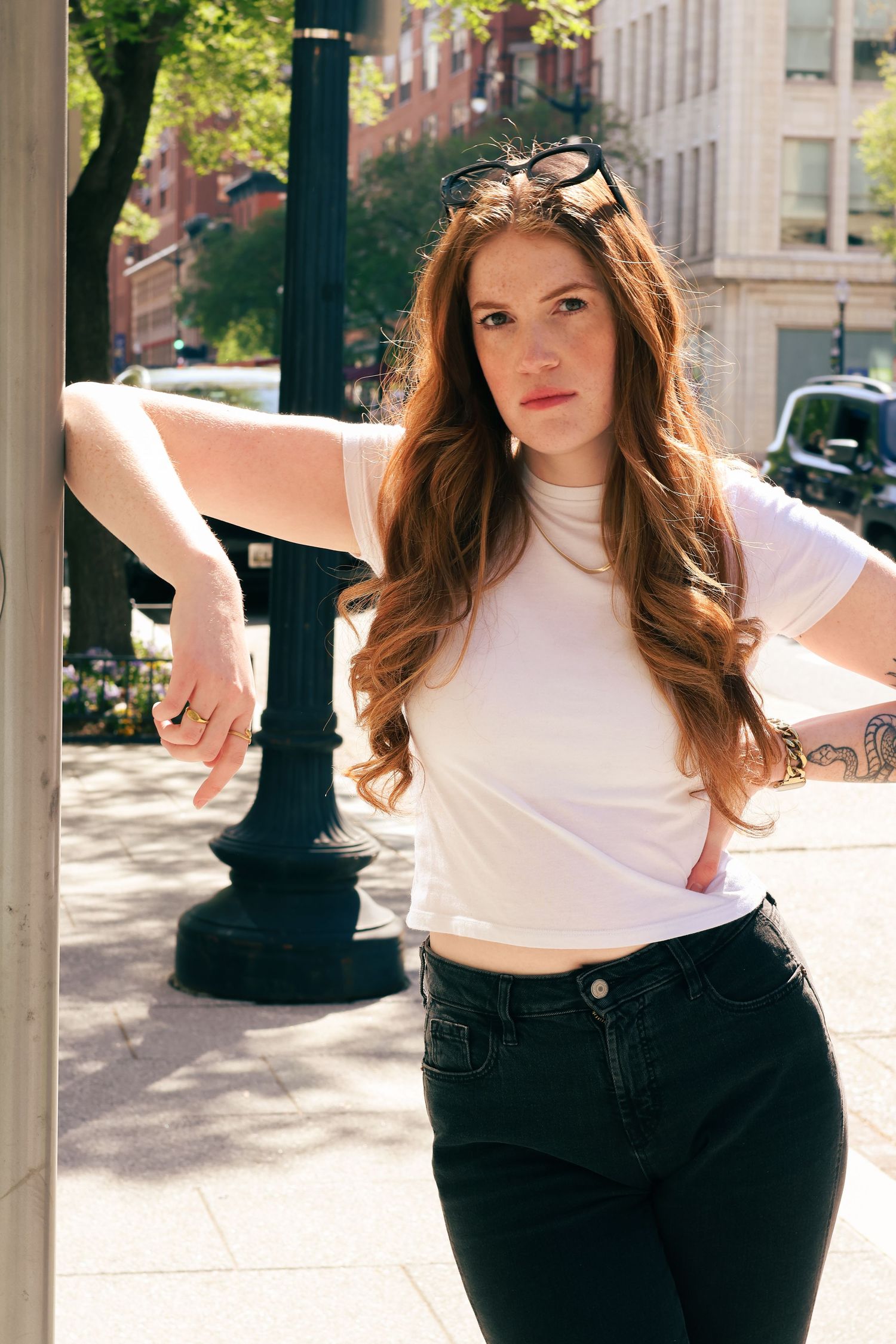 Redhead woman in white crop top and black jeans leaning on pole, sunglasses on head, tattoo on arm, city street.