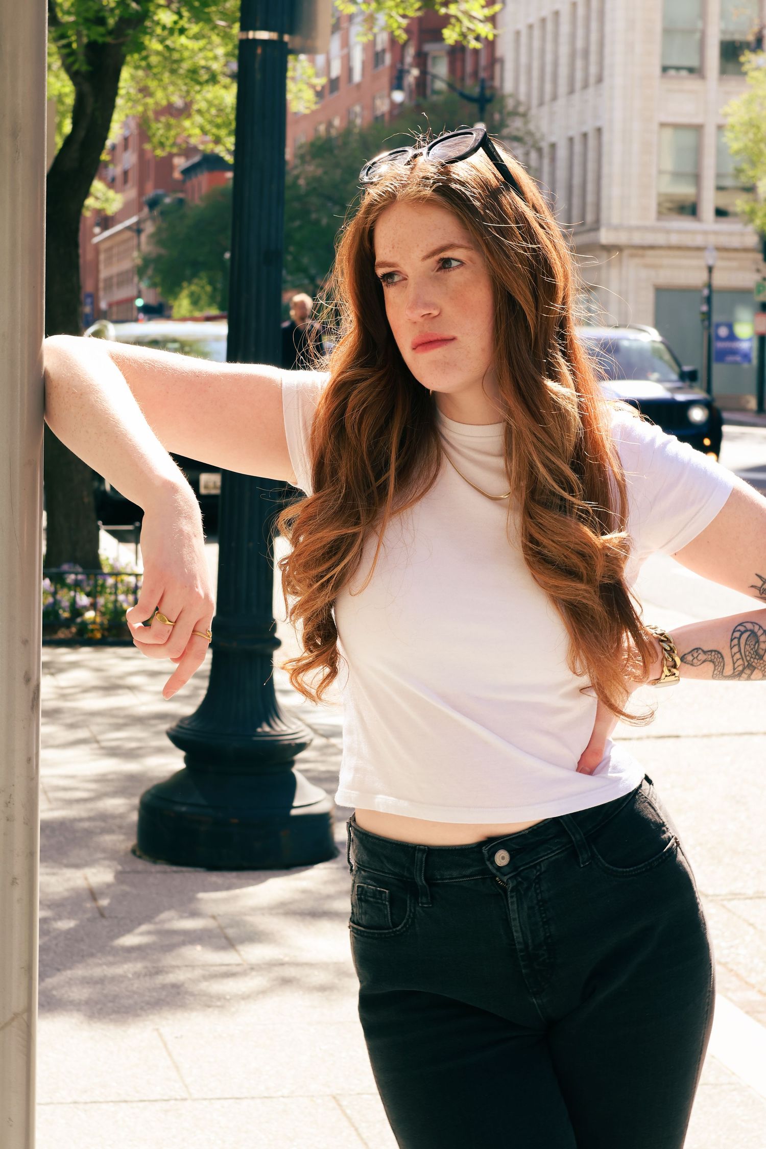 Red-haired tattooed woman in white crop top leans on lamppost on sunny urban street.