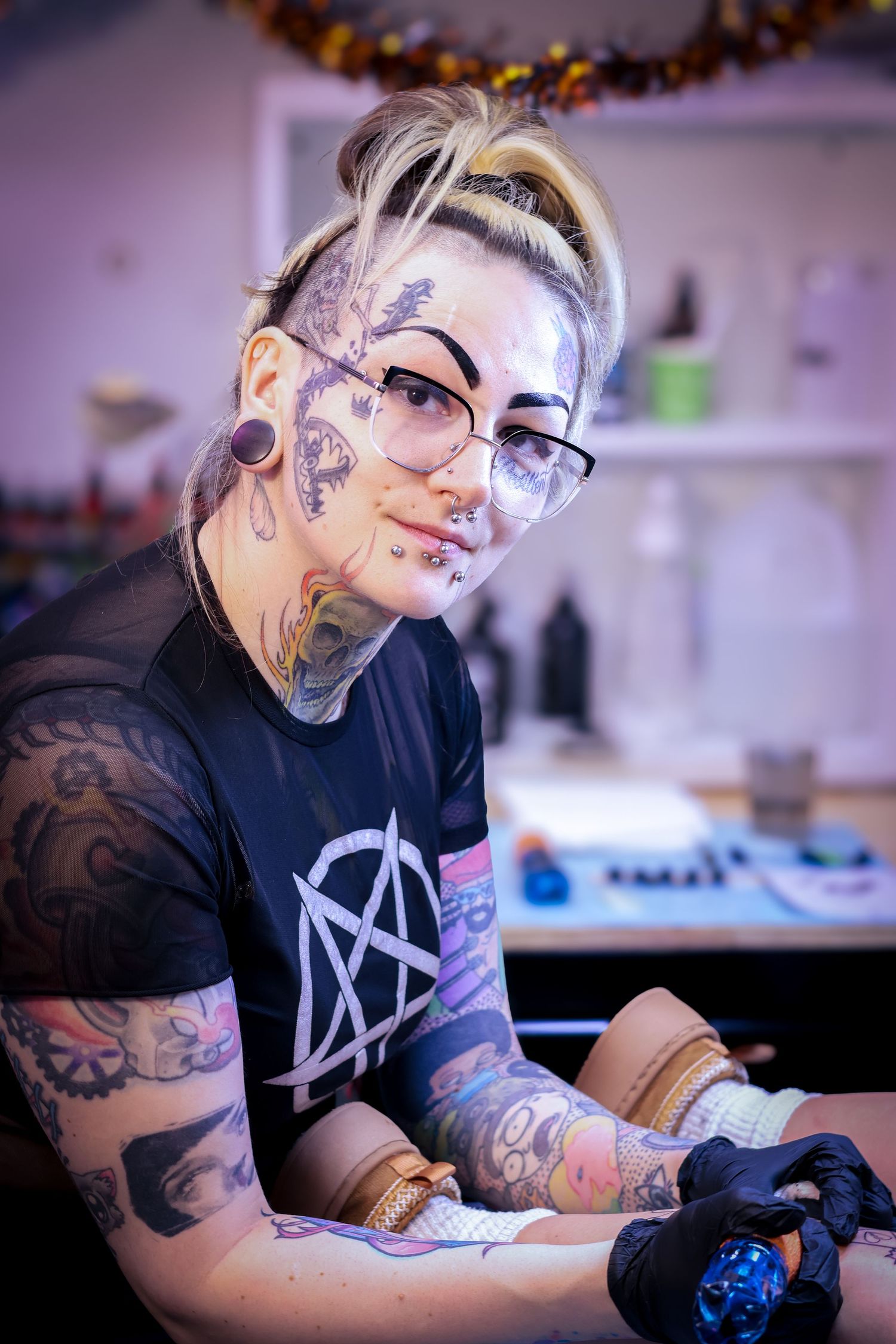 A tattoo artist wearing black gloves works diligently at their station in a studio with decorative lights overhead.