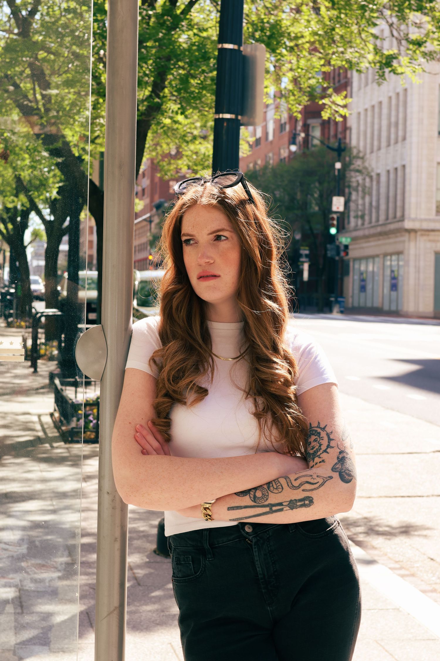 Young woman with long wavy red hair, tattoos, and crossed arms leans against a pole on a sunny city street.
