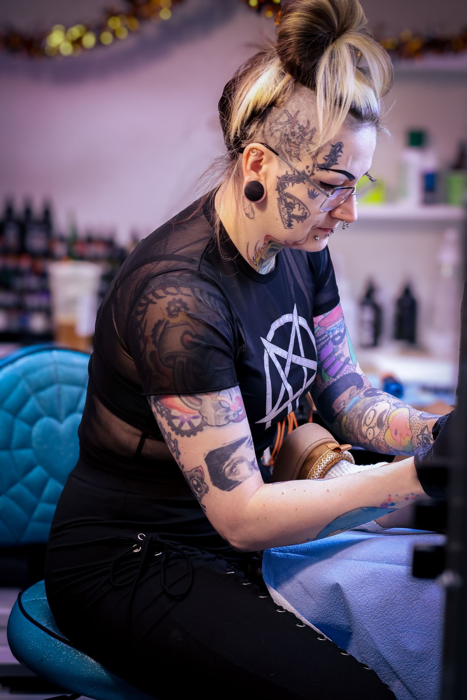 Tattoo artist in black shirt working intently at a station with blue chair and decorative lighting.