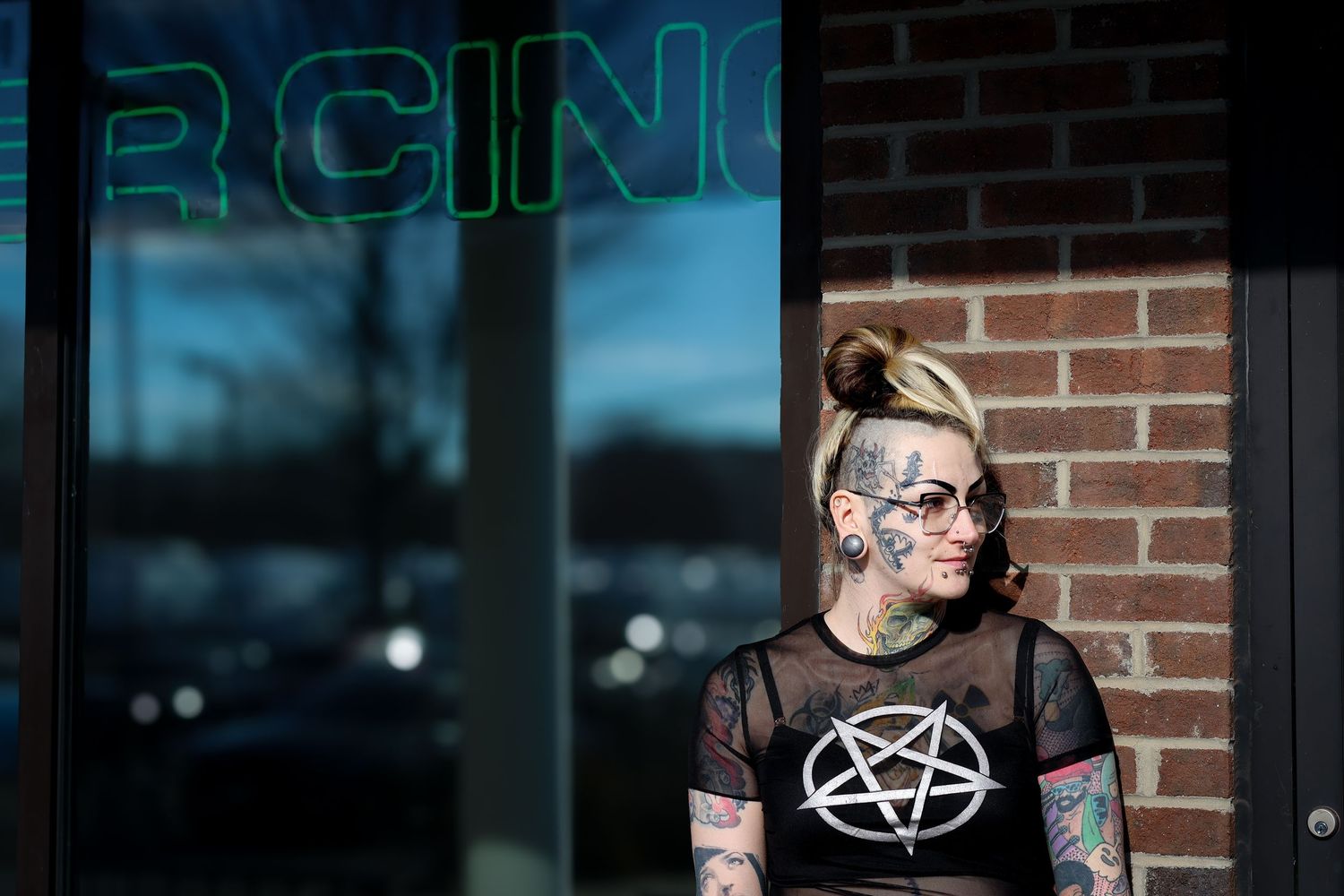 A person in a black pentagram shirt stands against a brick wall with neon signage visible in the background.