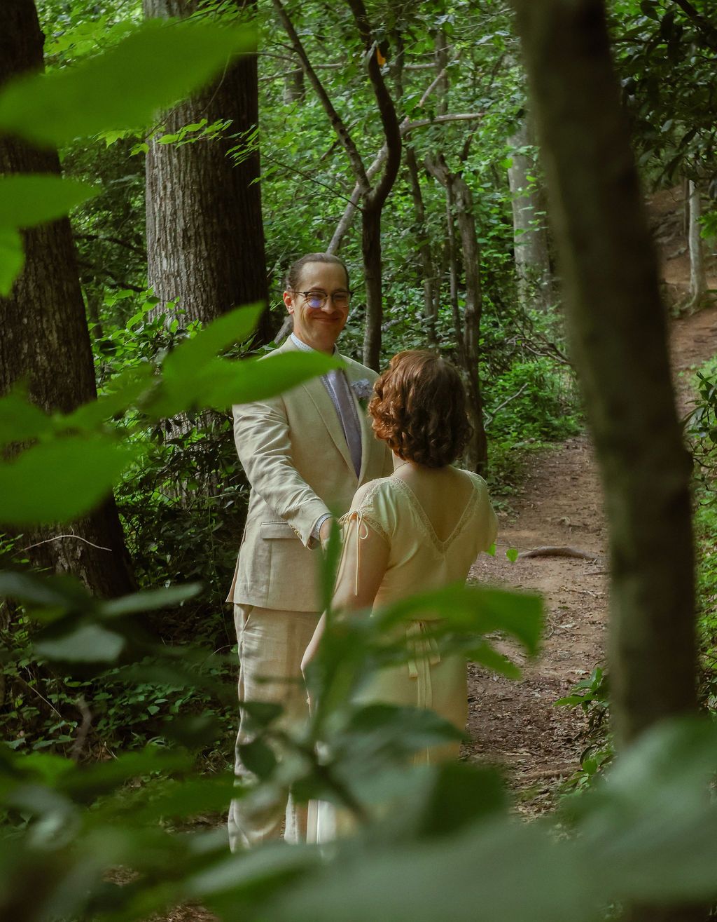 Groom in a tan suit and bride in a cream dress smile at each other on a leafy forest trail.