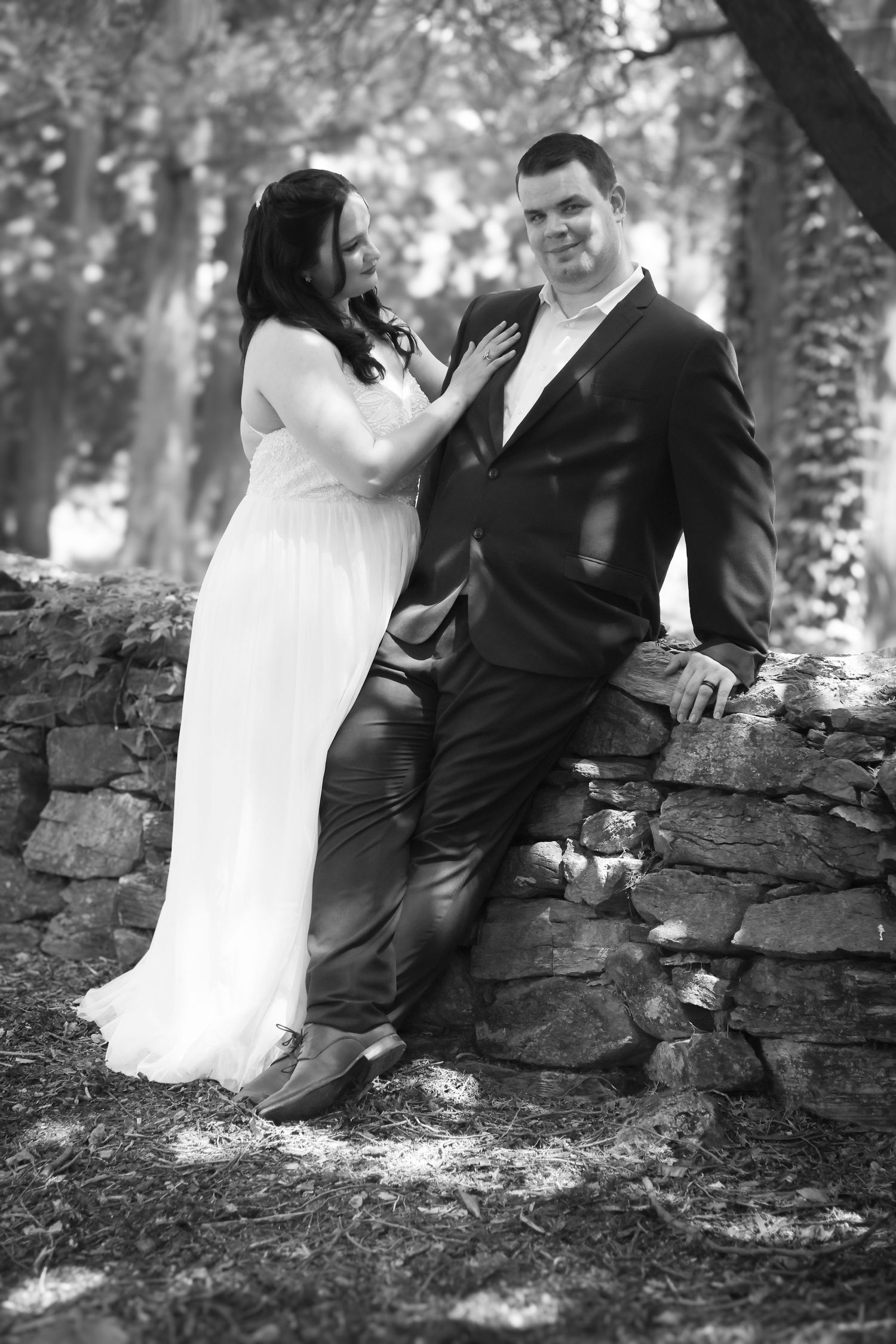 A romantic black and white wedding photo of a couple posing against a rustic stone wall in a wooded setting.