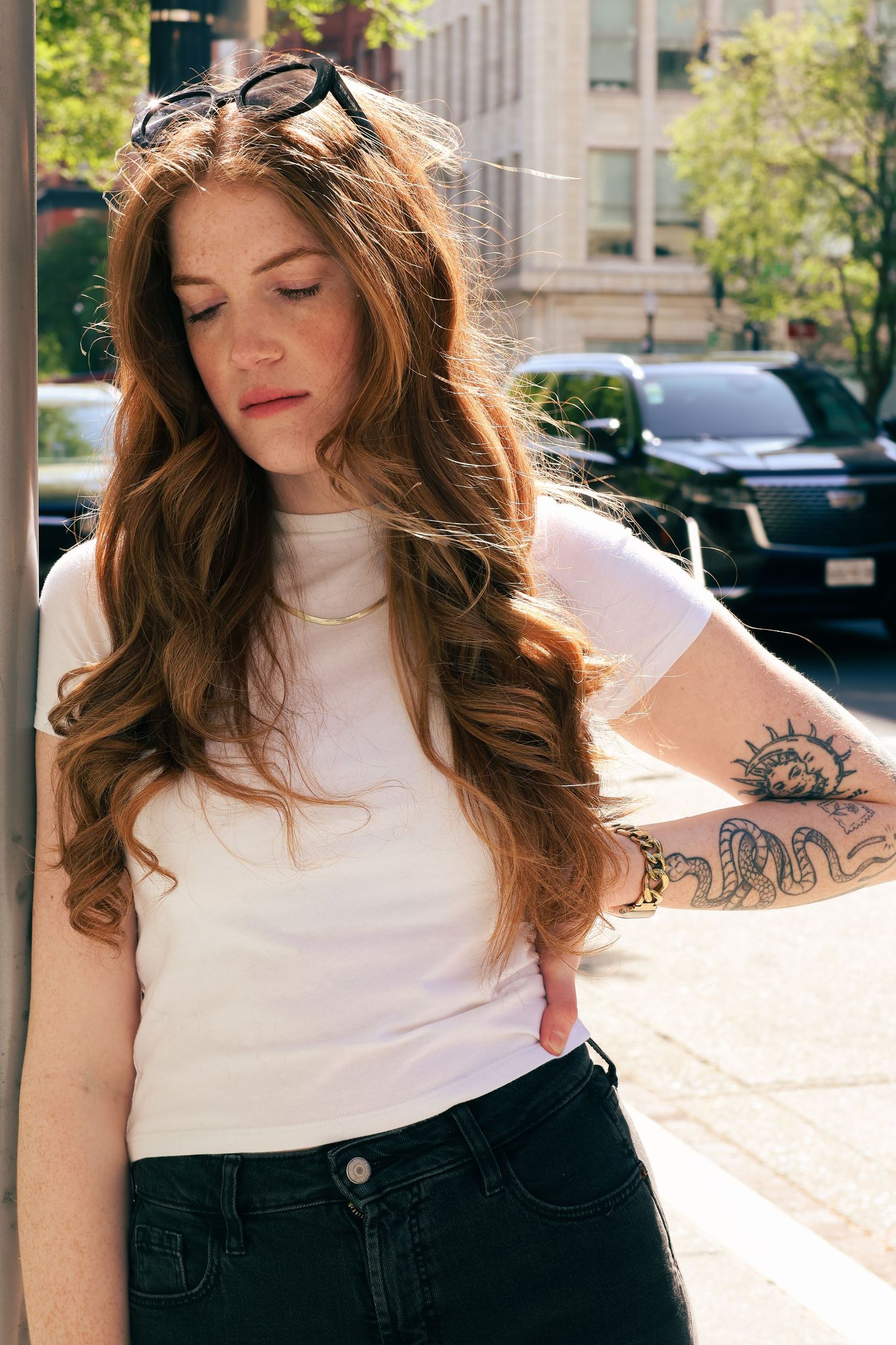 Young woman with long auburn hair and arm tattoo wearing white tee and black jeans outdoors.