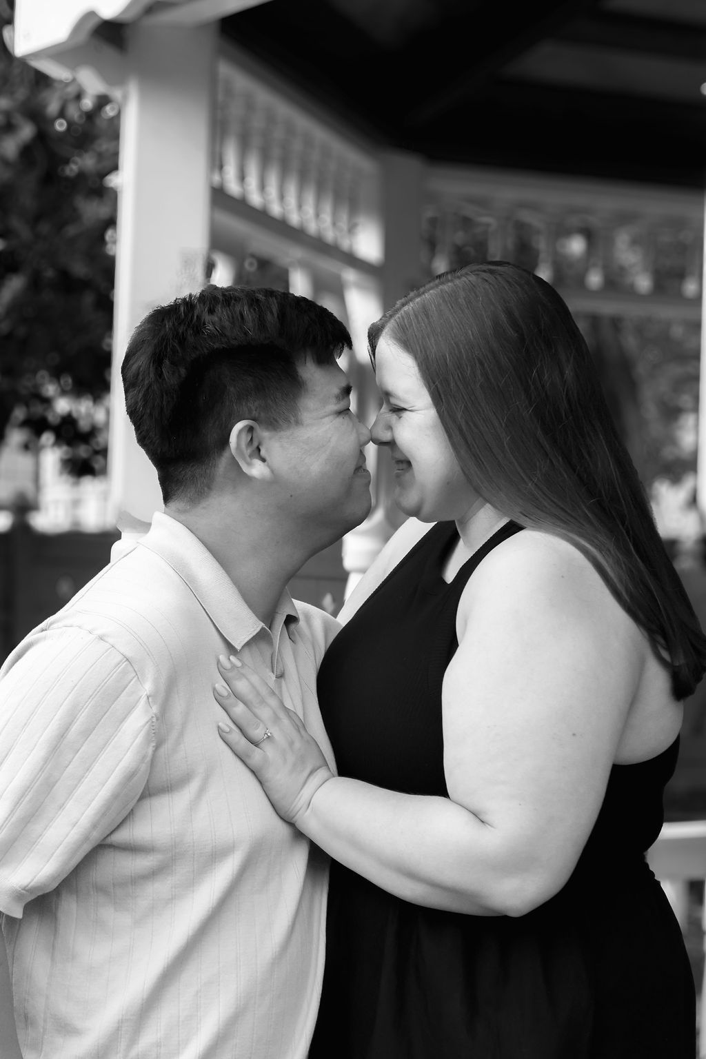 Black and white engagement portrait of a couple touching noses and smiling affectionately in front of a white gazebo.