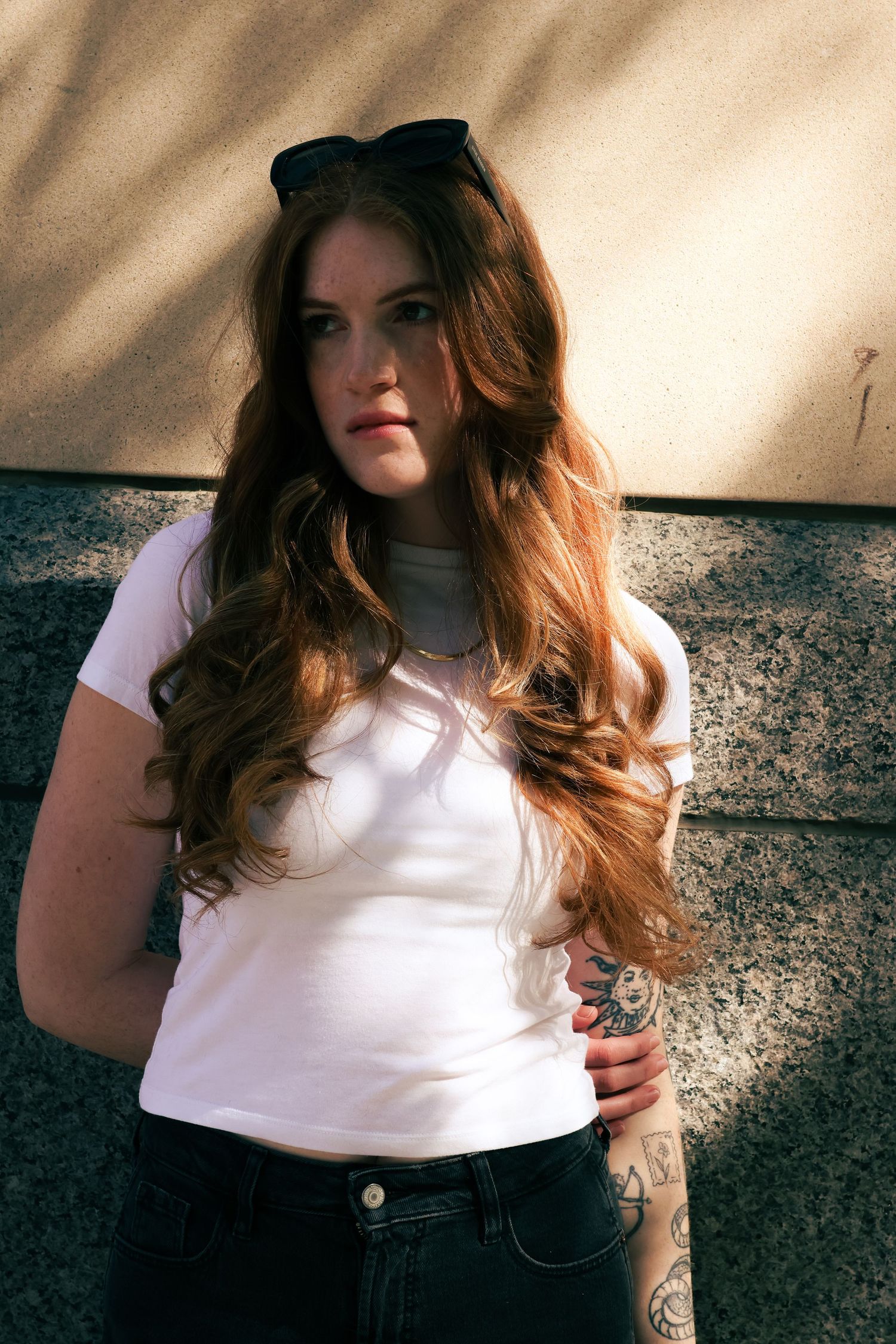 Young woman with long auburn hair in white tee and black jeans poses against stone wall outdoors.