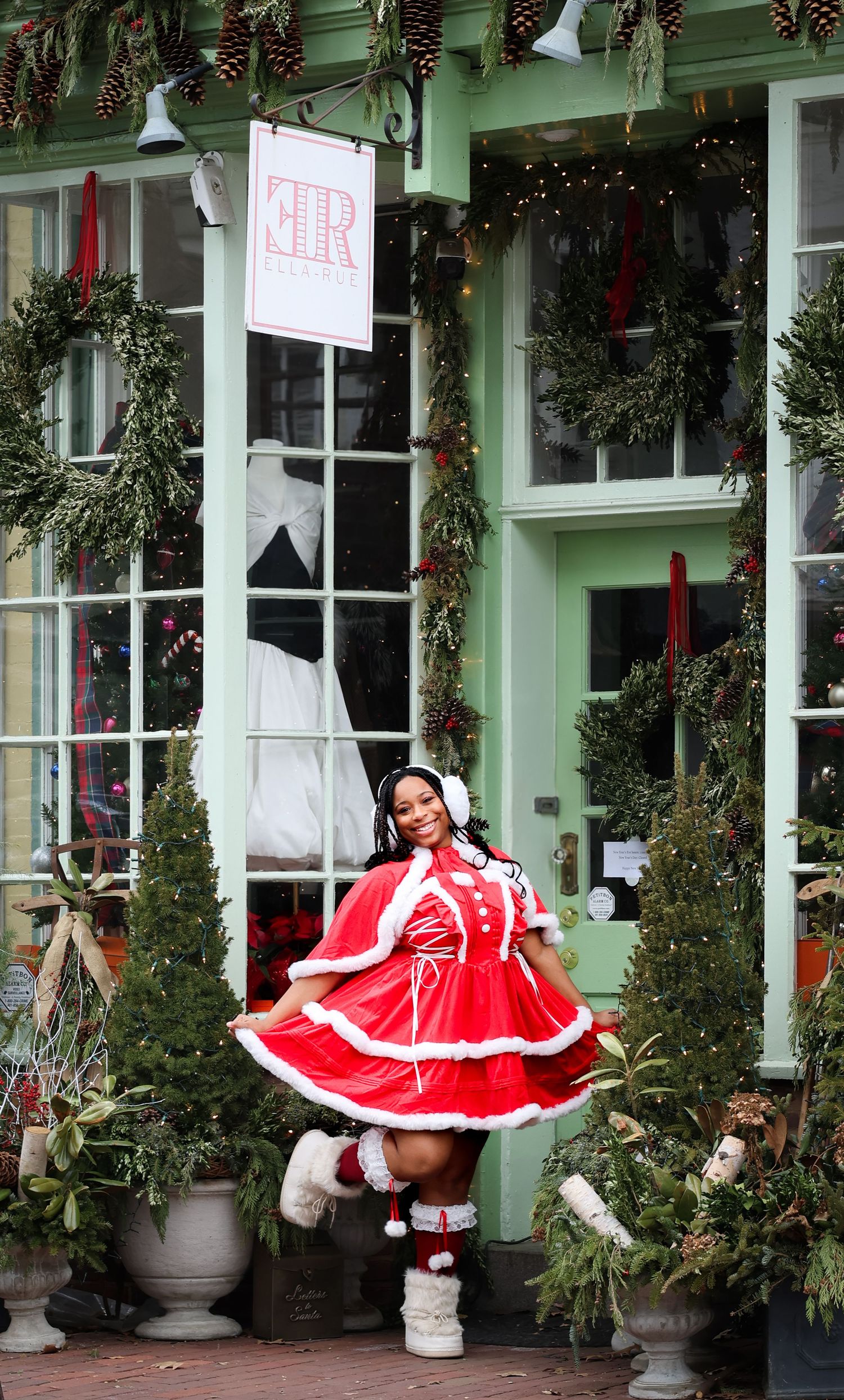 Woman in red and white Mrs. Claus costume posing in front of a festively decorated storefront with wreaths.