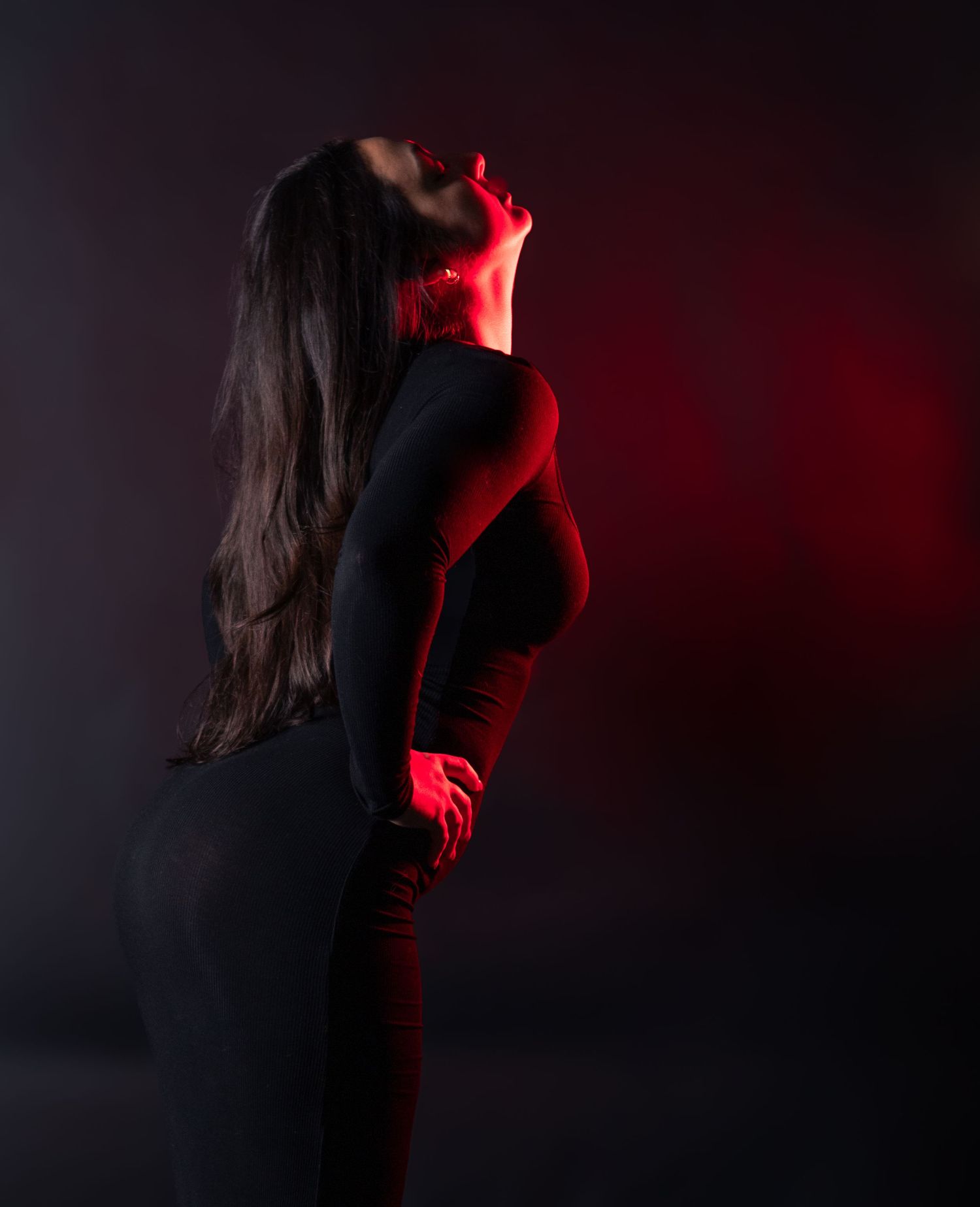 Woman in black outfit posed dramatically with red lighting against dark background.