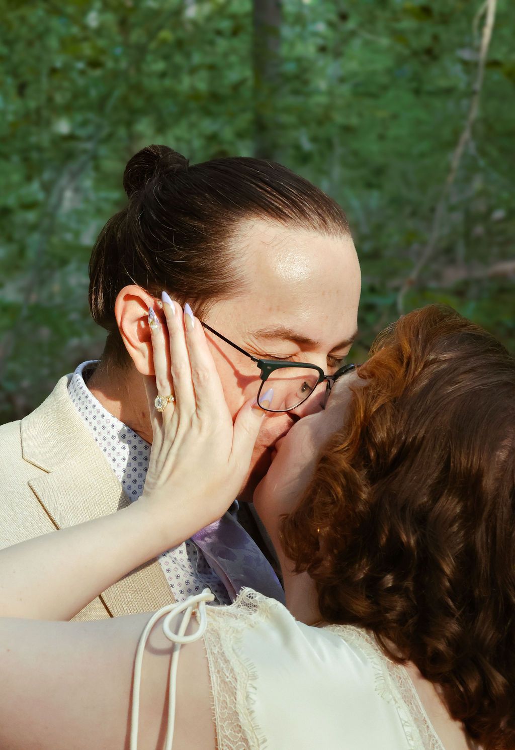 Couple sharing a kiss in the woods, her hand with a ring on his cheek, he wears glasses and a tan suit.