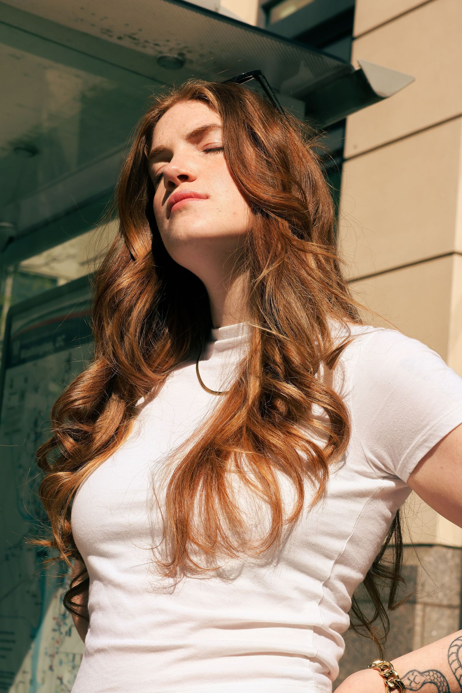 Young woman with long wavy auburn hair wearing a white t-shirt, eyes closed in sunlight outdoors.