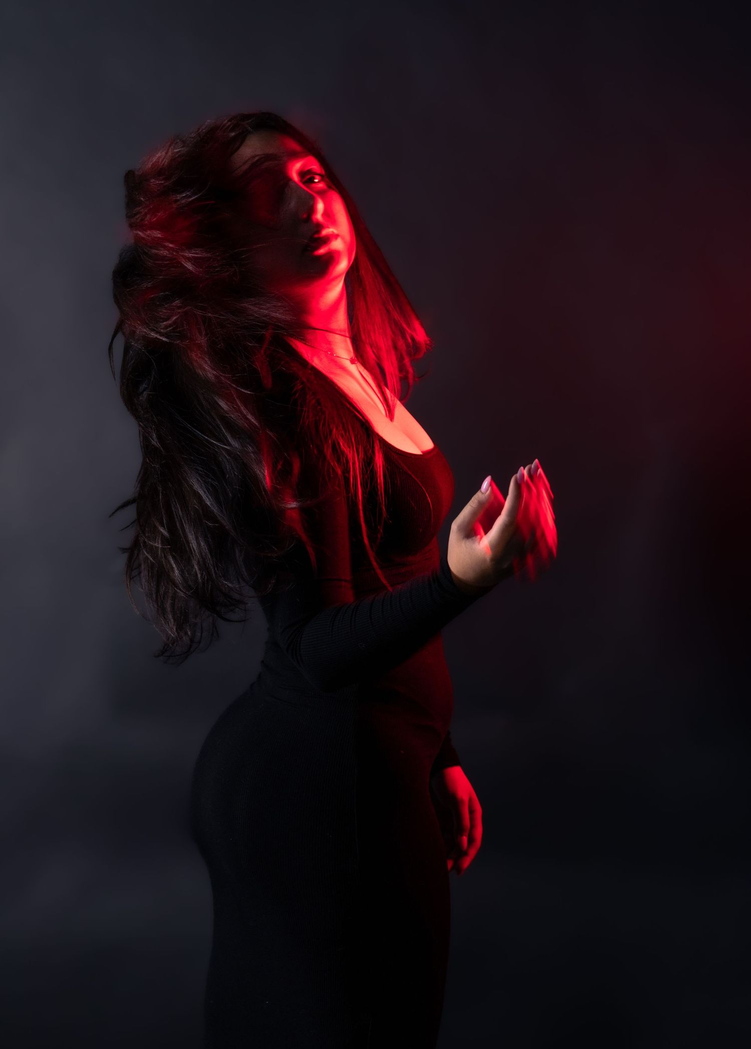 Woman in black dress with hair flowing under intense red light against dark background.
