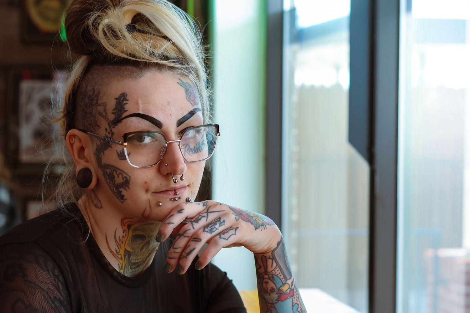 Person with facial tattoos and glasses gazes thoughtfully by window in natural lighting.
