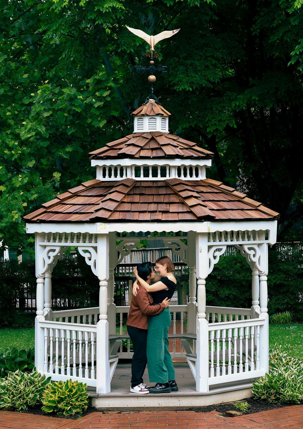 Couple embracing inside a white wooden gazebo with a cedar shingle roof and bird weathervane, surrounded by green trees.