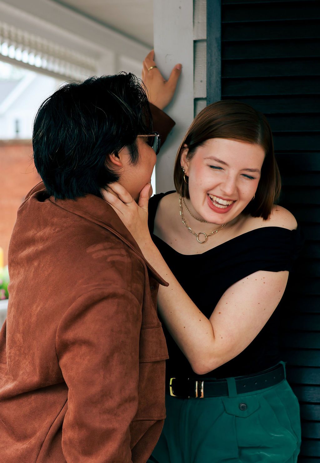 Two friends laugh together on a porch as a woman in a black top and green pants smiles while a person in a brown jacket leans in.