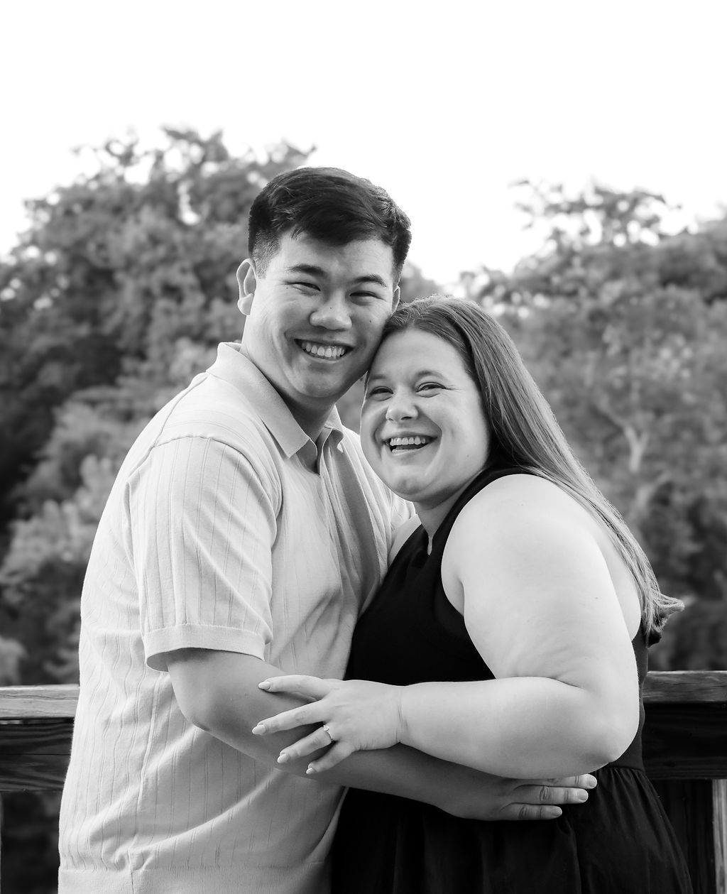 Black and white portrait of a joyful couple laughing and embracing outdoors, with trees softly blurred in the background.
