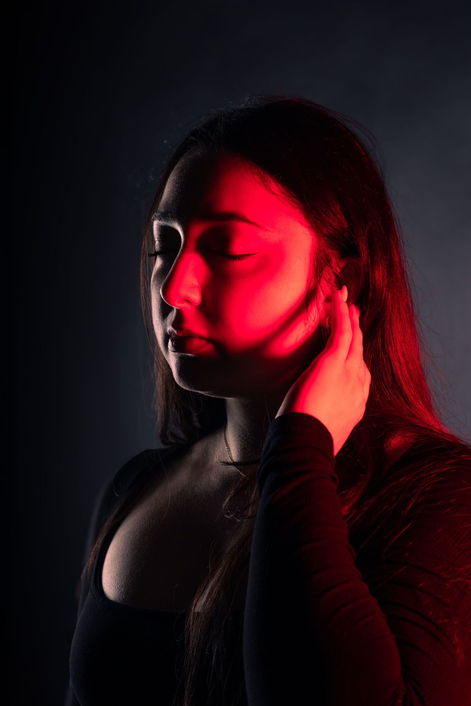 Woman with closed eyes touched by red light in moody dark studio portrait.
