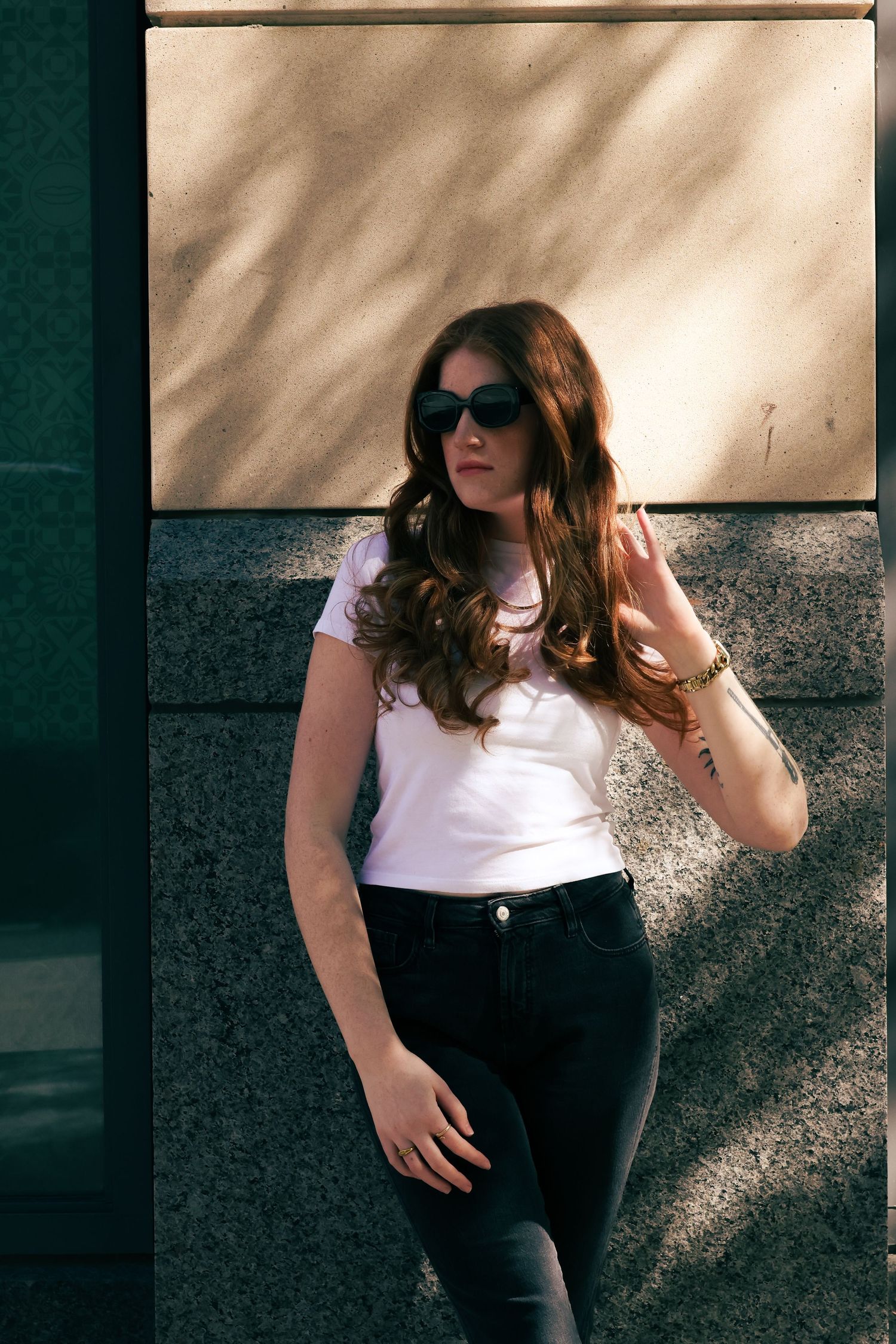 Young woman with long wavy hair wearing sunglasses, white t-shirt, and black jeans posing against a stone wall outdoors.
