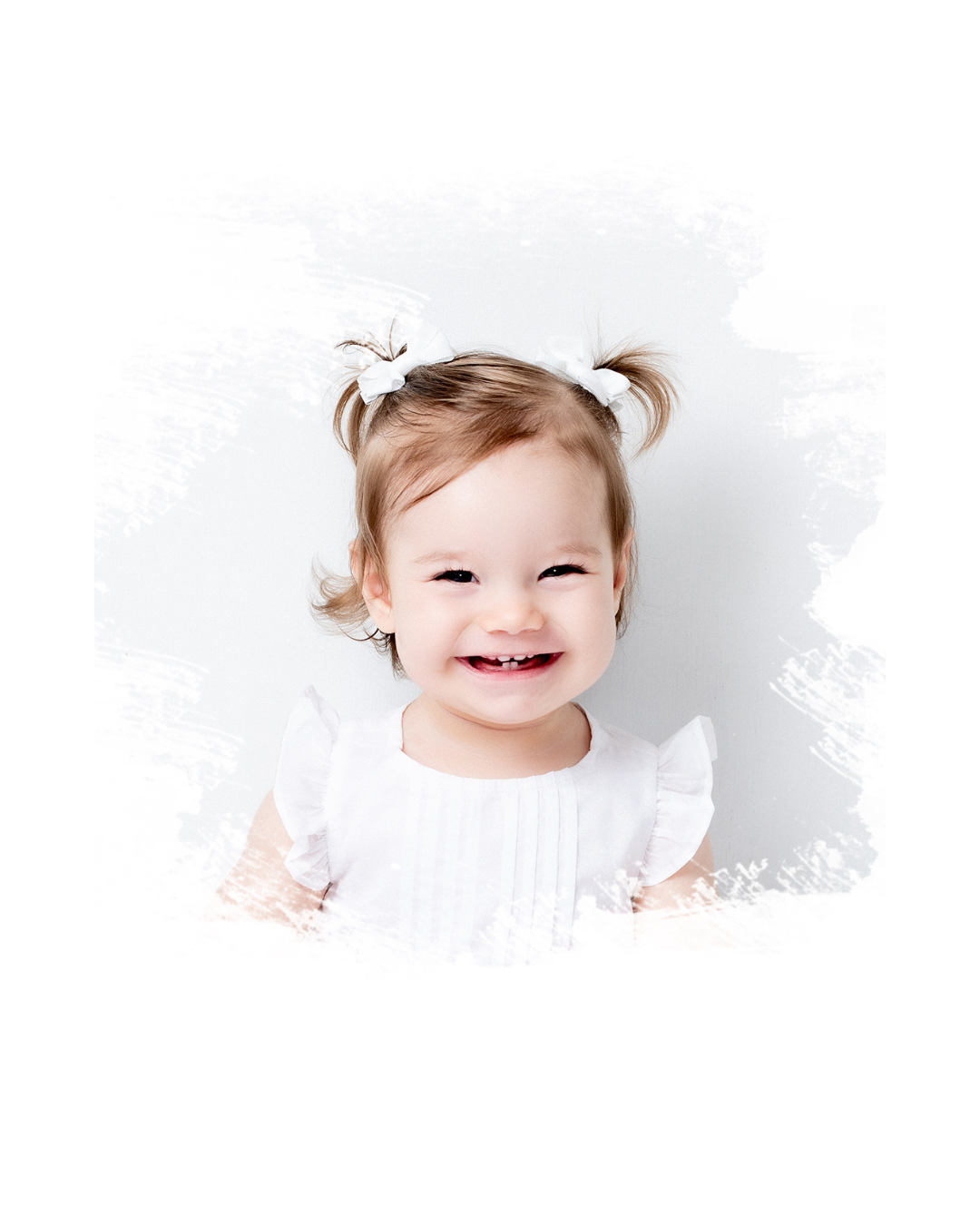 A cheerful toddler in a white dress smiles brightly against a white background with their hair in small pigtails.