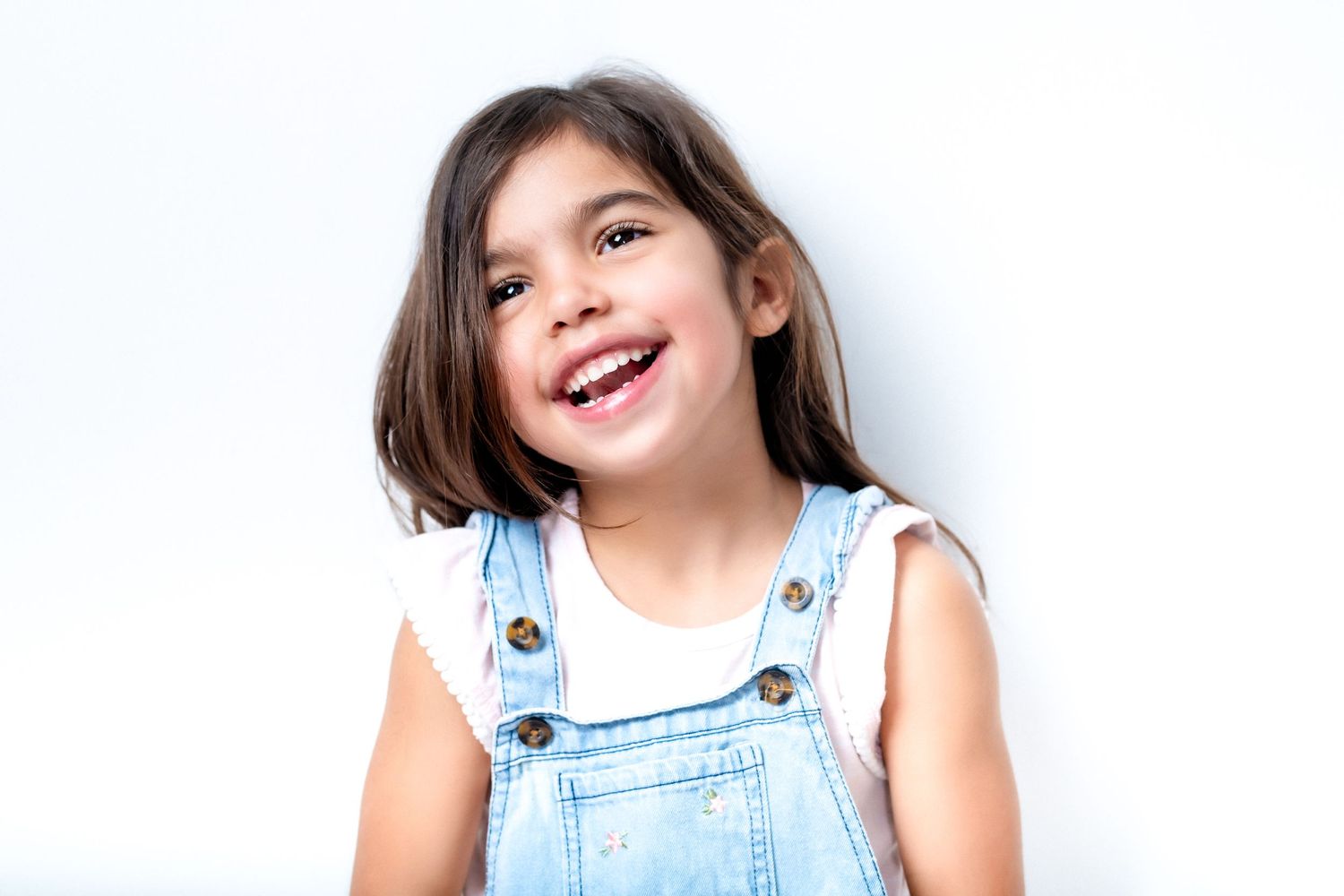 A joyful child in denim overalls smiles authentically against a white background.