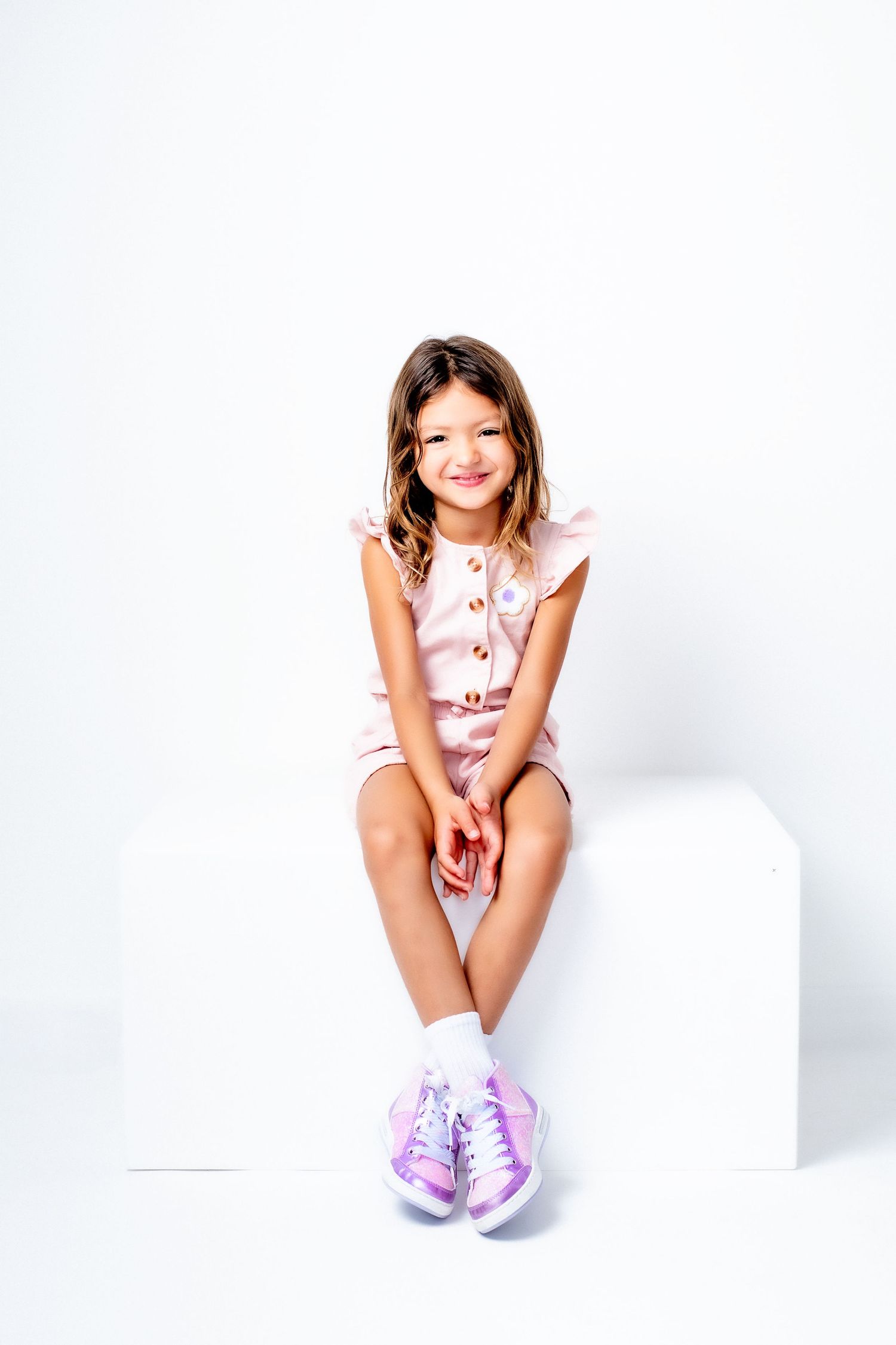 A person in pink clothing and purple sneakers sits on a white surface against a bright background.