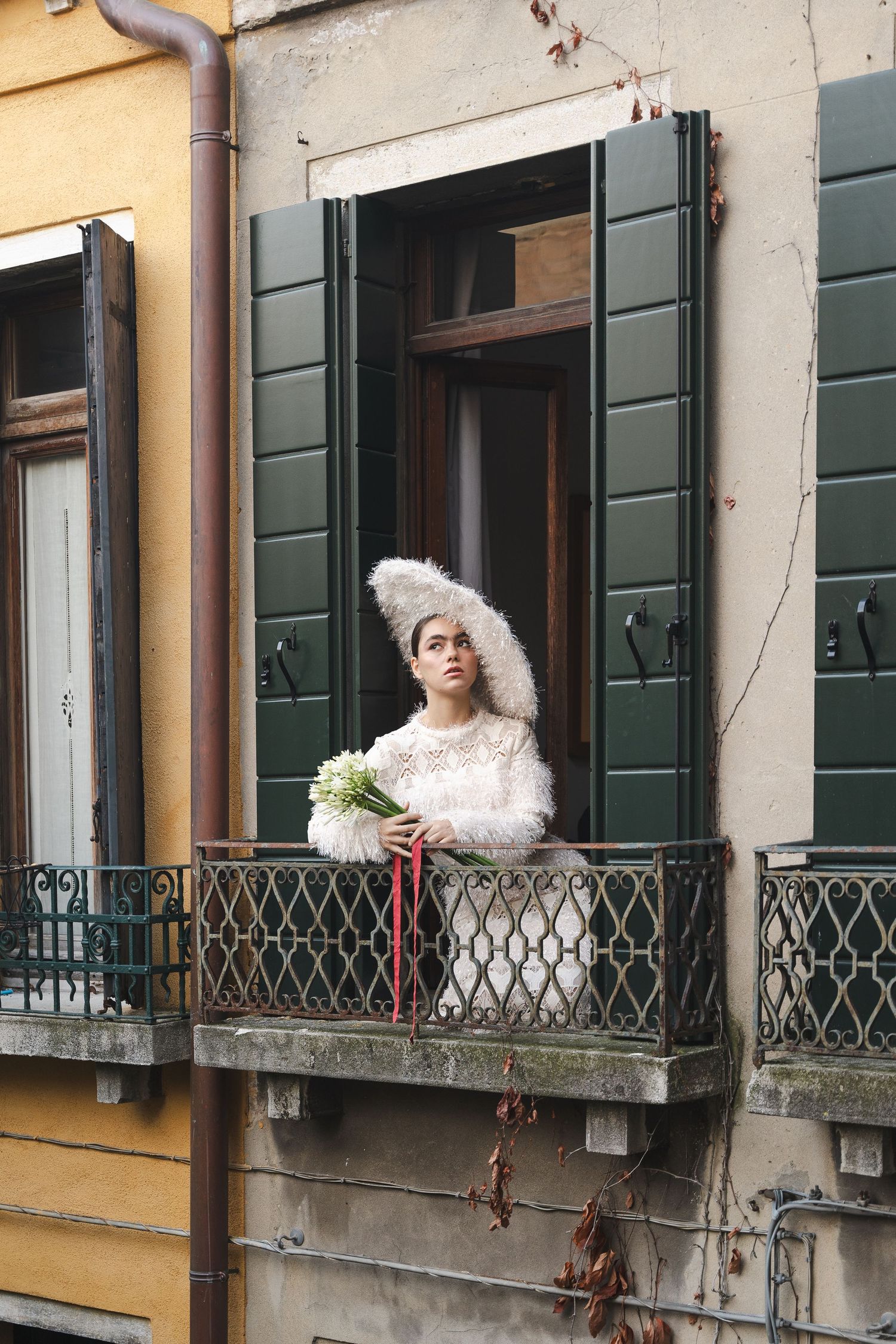 Fashion editorial on Venetian balcony featuring white feathered dress and classic shutters.