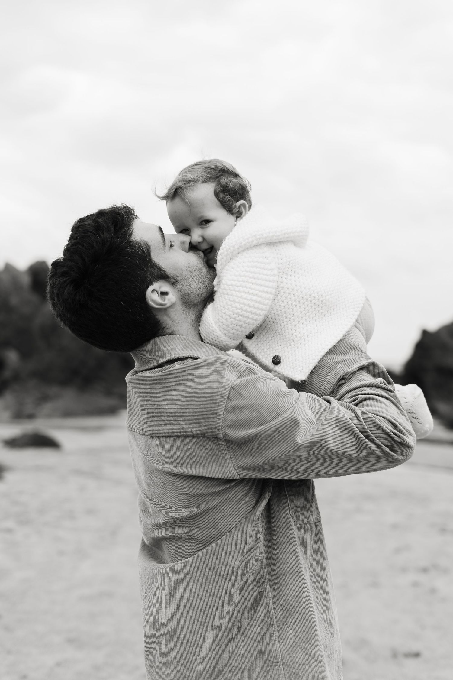 Black and white photo capturing tender moment between parent and baby at beach.