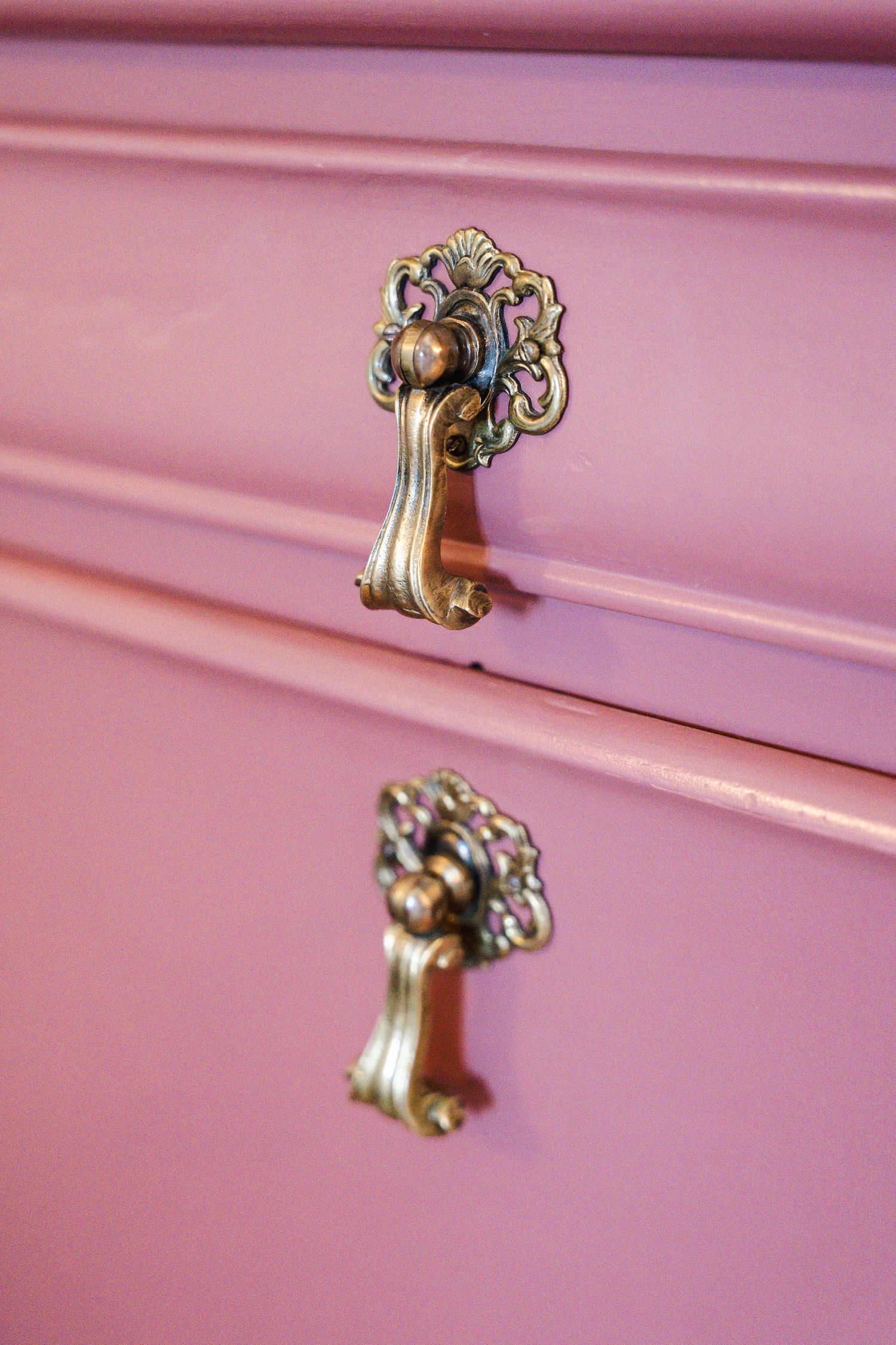 Ornate brass drawer pulls on pink painted furniture with decorative details.
