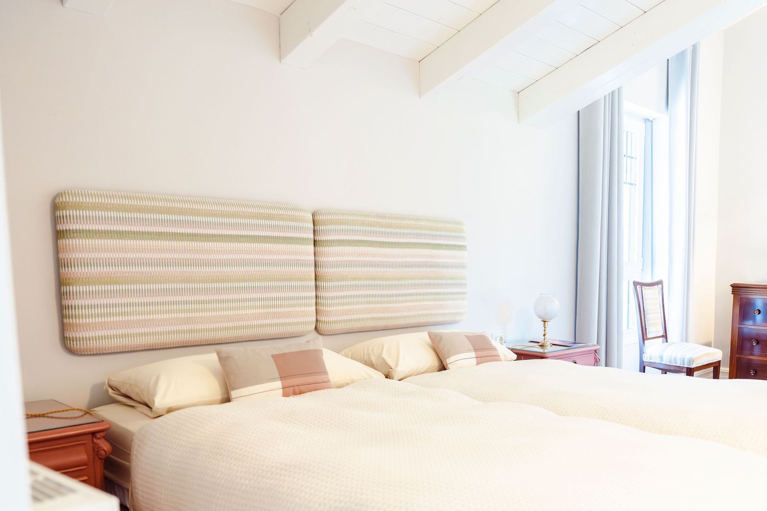 Airy bedroom with striped upholstered headboard, neutral bedding, and exposed ceiling beams.