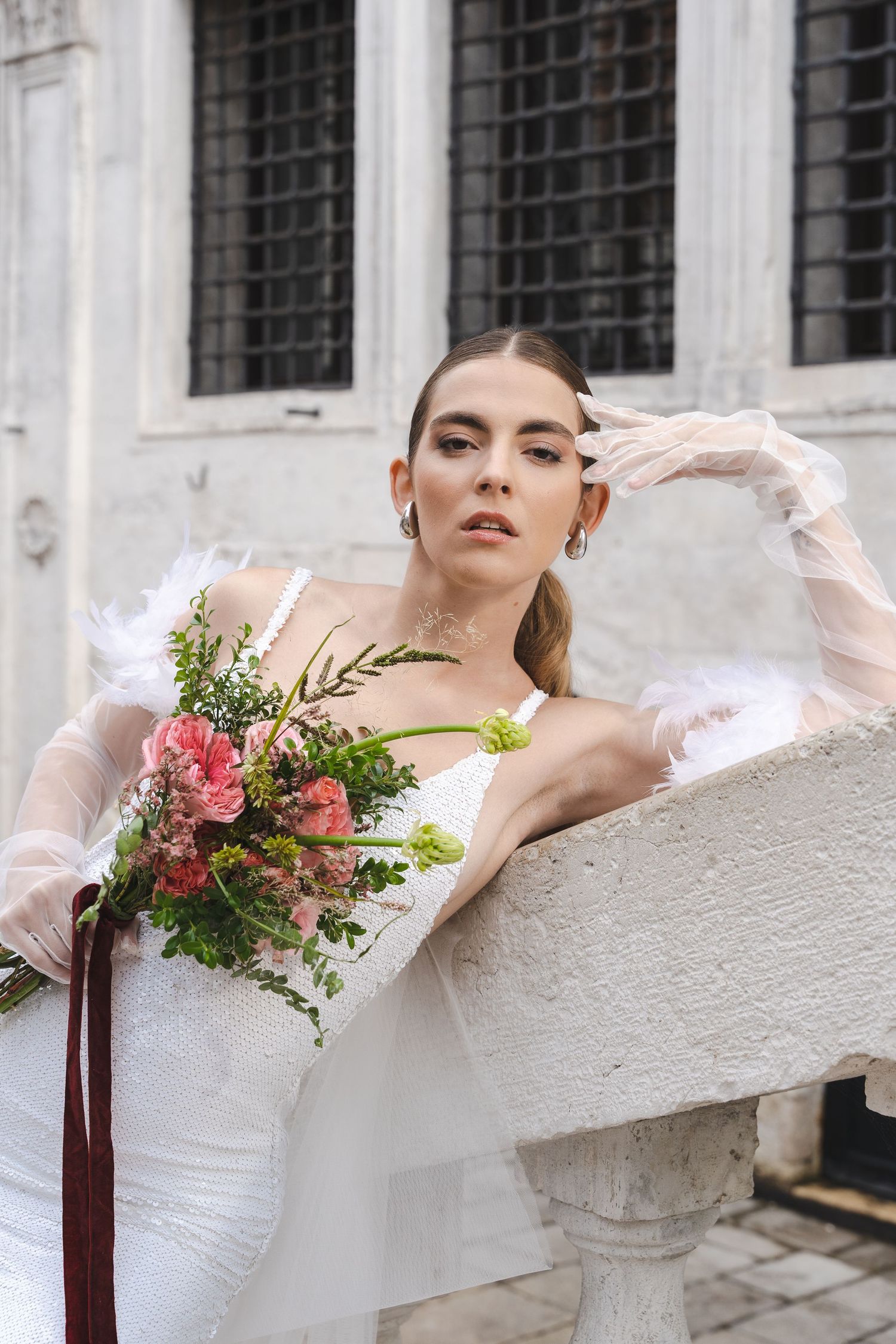 A romantic bridal portrait features a flowing white dress, delicate gloves, and a vibrant wildflower bouquet against stone architecture.