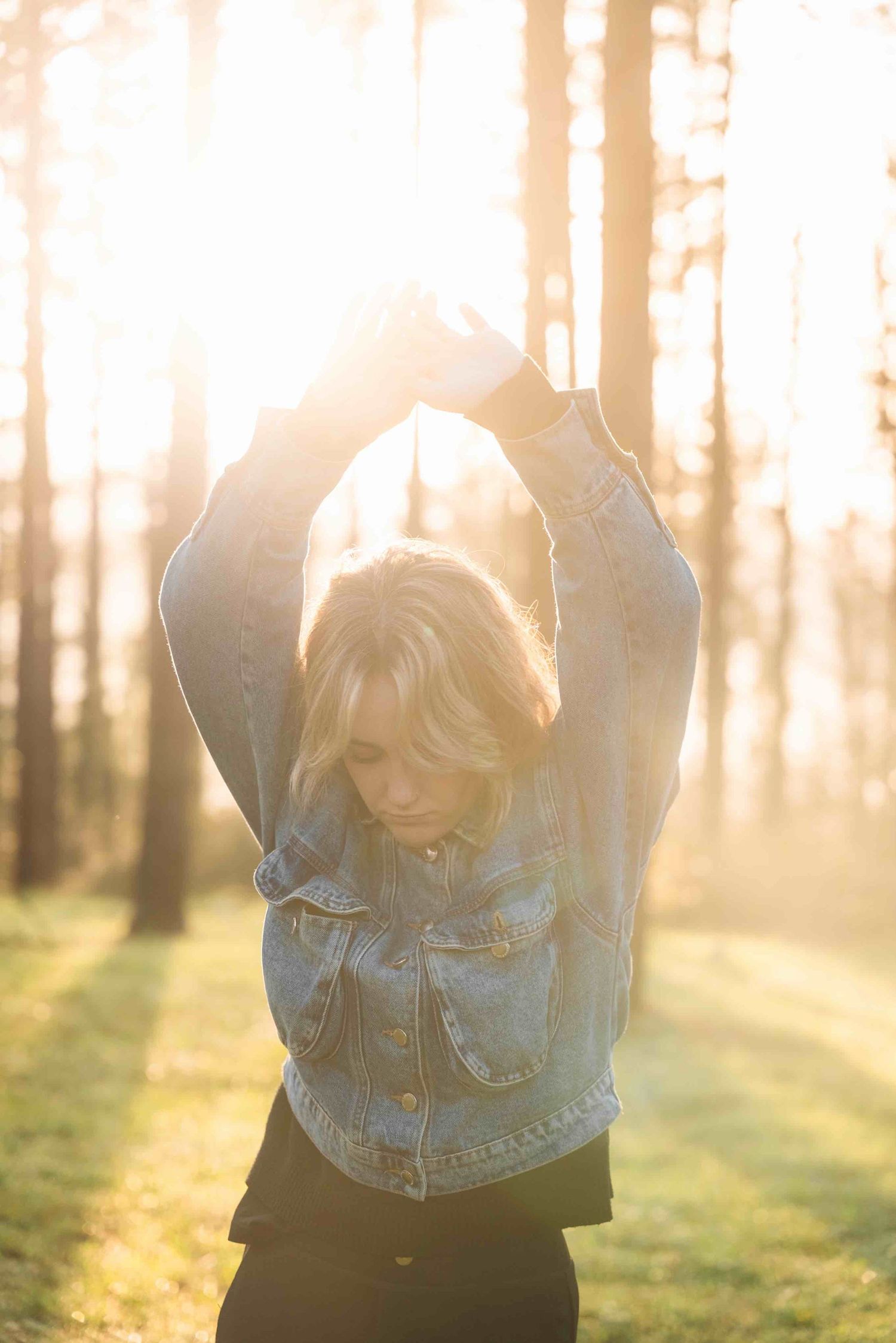 Person stretches arms overhead in denim jacket during golden hour in forest setting.