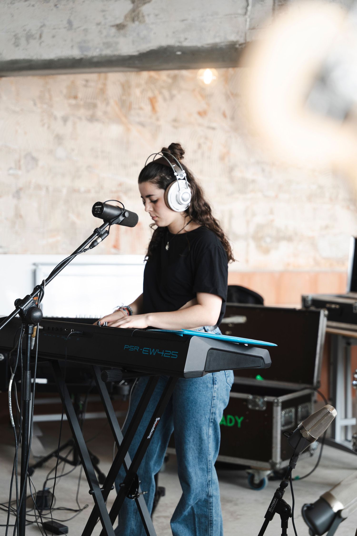 A musician performs at a keyboard setup with headphones and microphone in an industrial space.