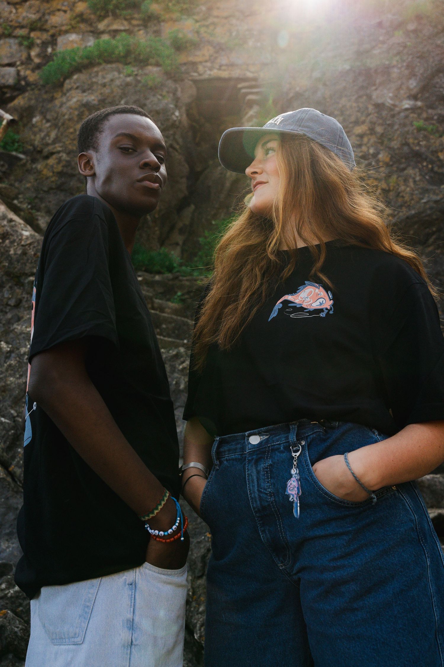 Two people in casual wear standing together against rocky backdrop in sunlight.