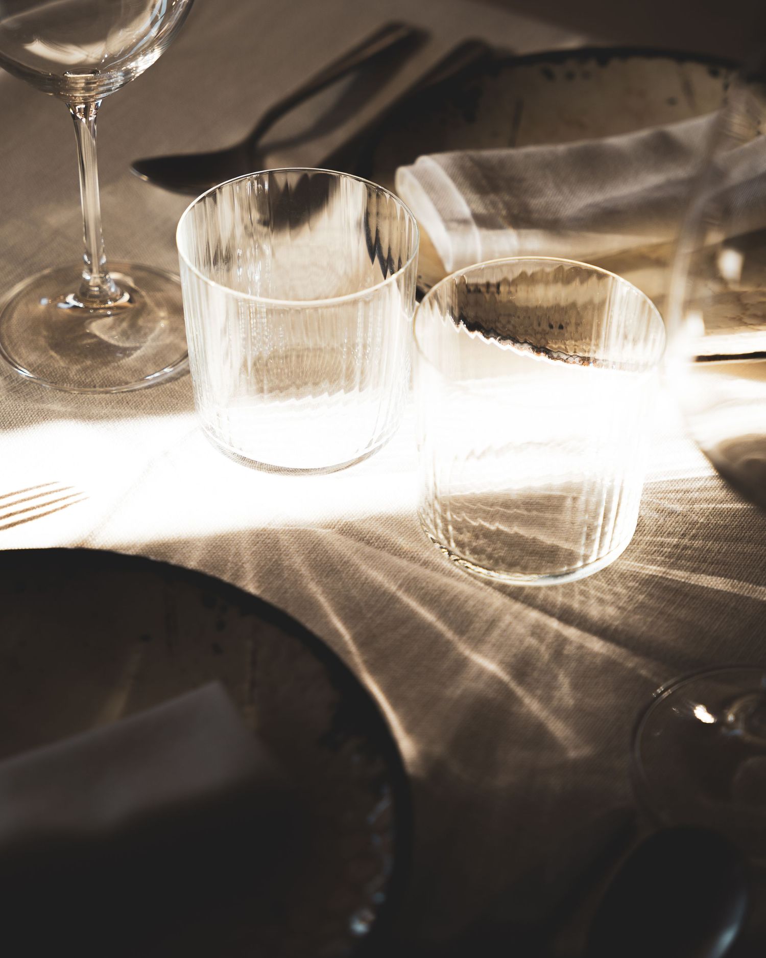 Elegant wine glasses and water tumblers catching afternoon light on a dark dining table.