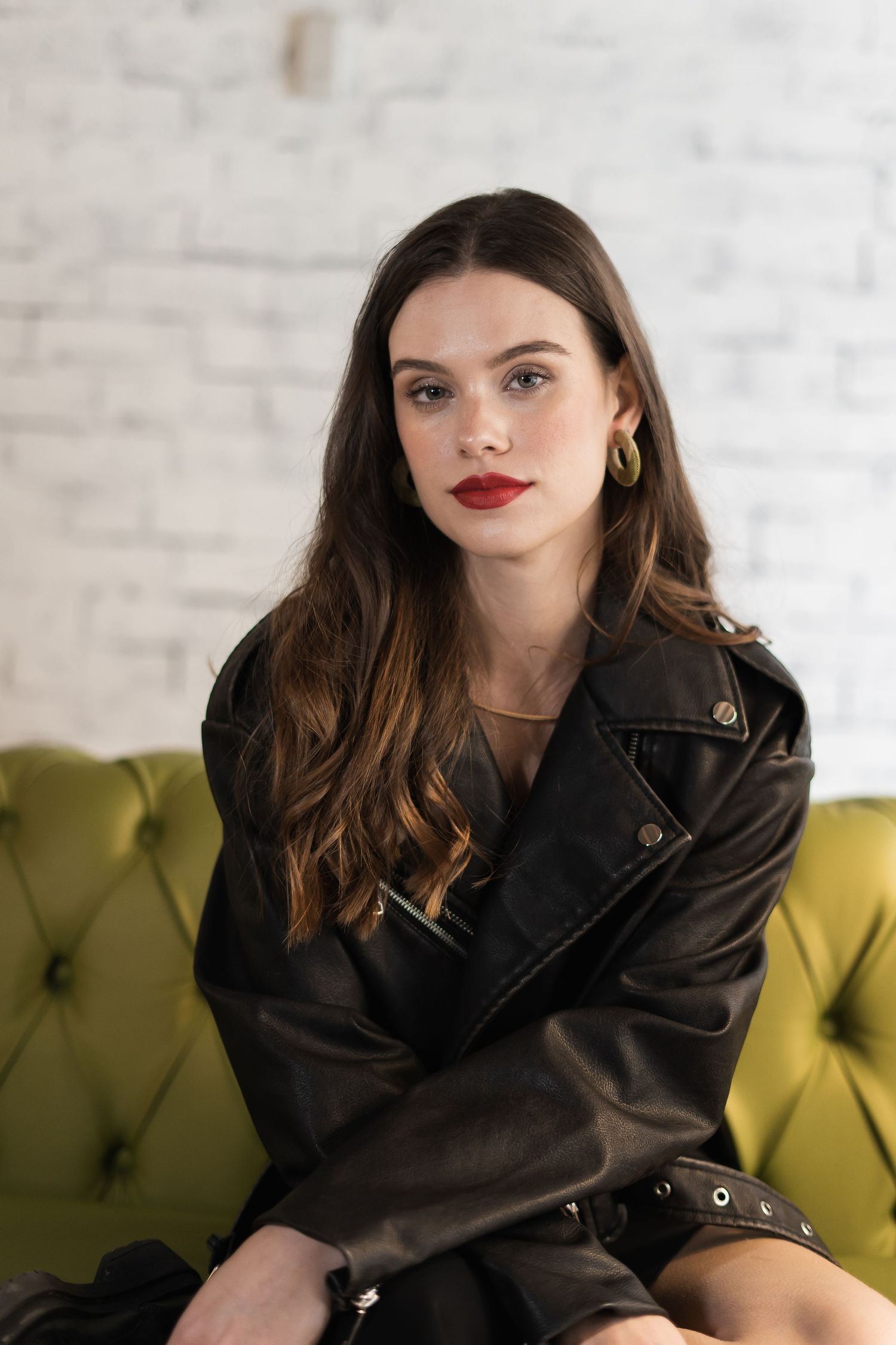 Fashion model wearing black leather jacket poses on yellow velvet couch against white brick wall.