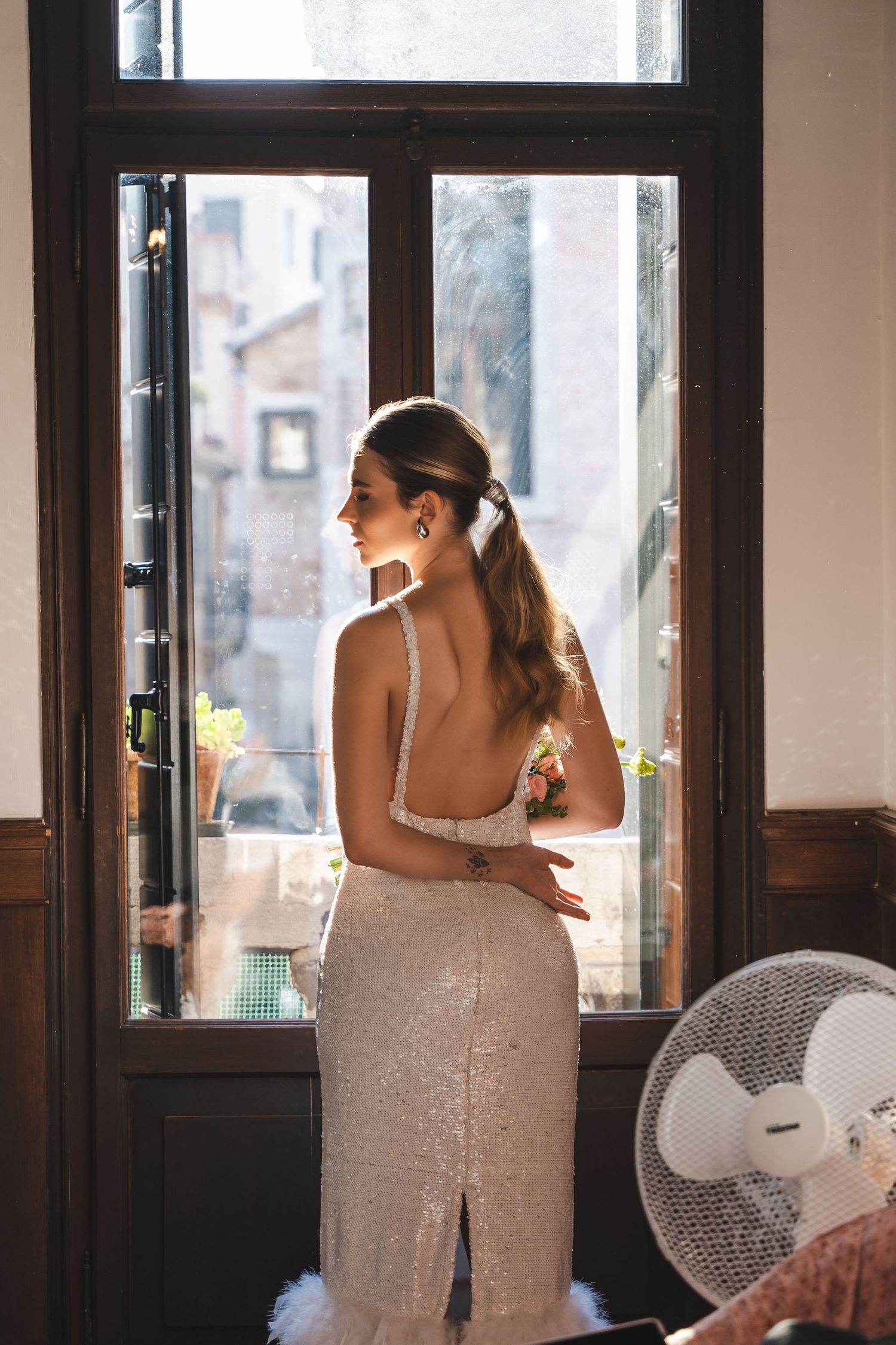 A woman in a shimmering formal dress stands by a window with her back turned, sunlight streaming through glass panels.