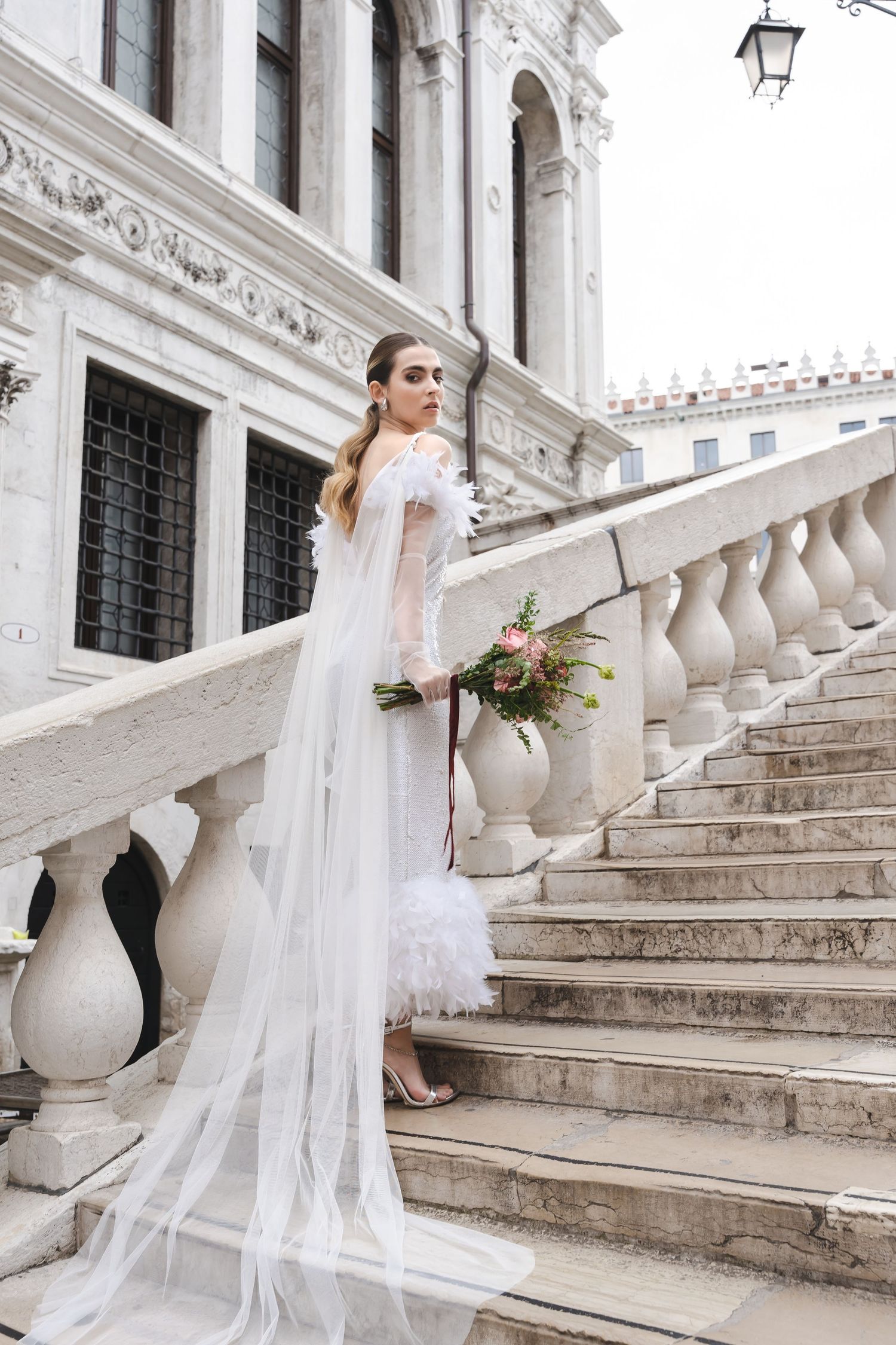 Bride in flowing white gown poses on grand marble staircase with classical architecture.