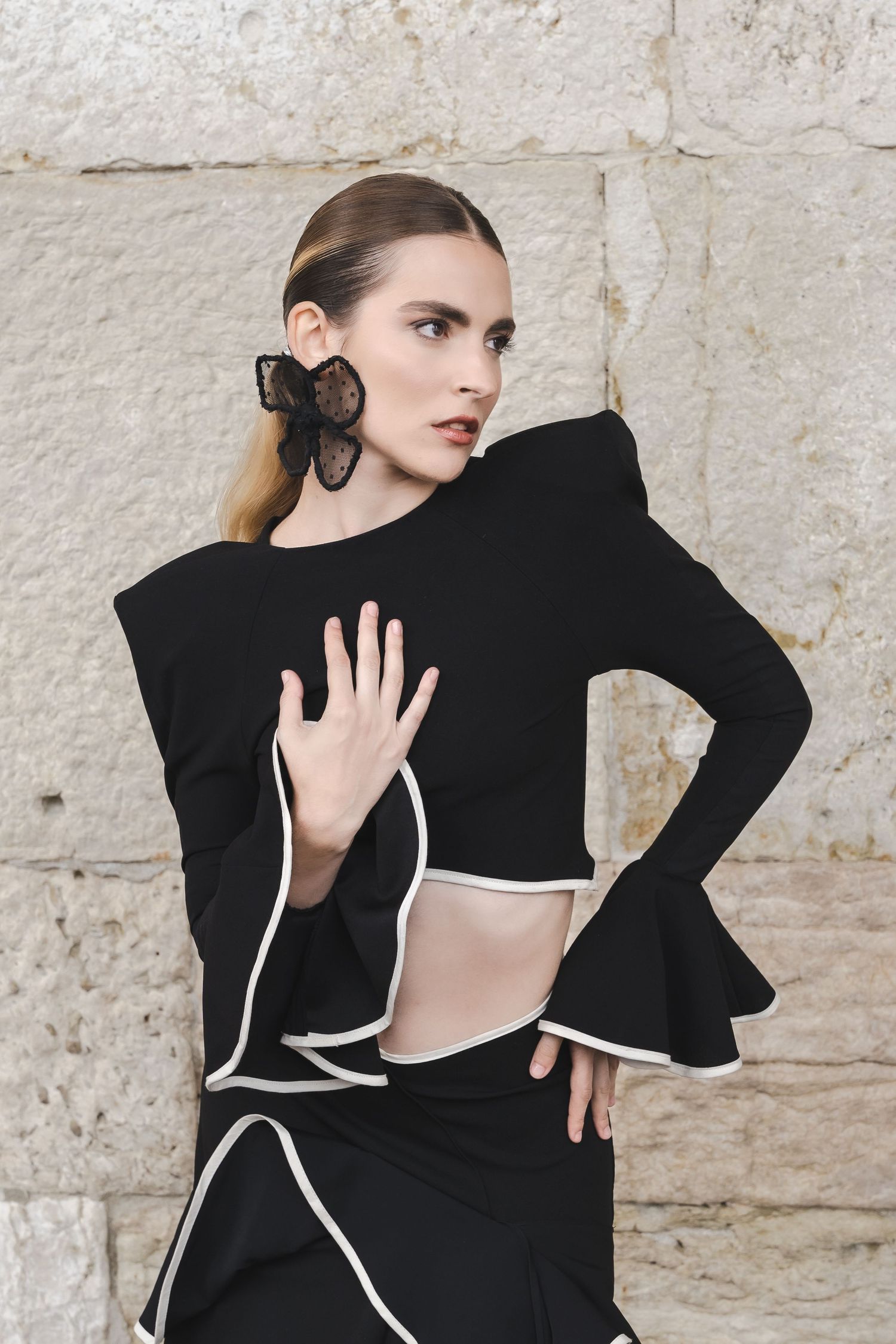 Fashion model wearing black cutout dress with ruffled sleeves poses against stone wall.
