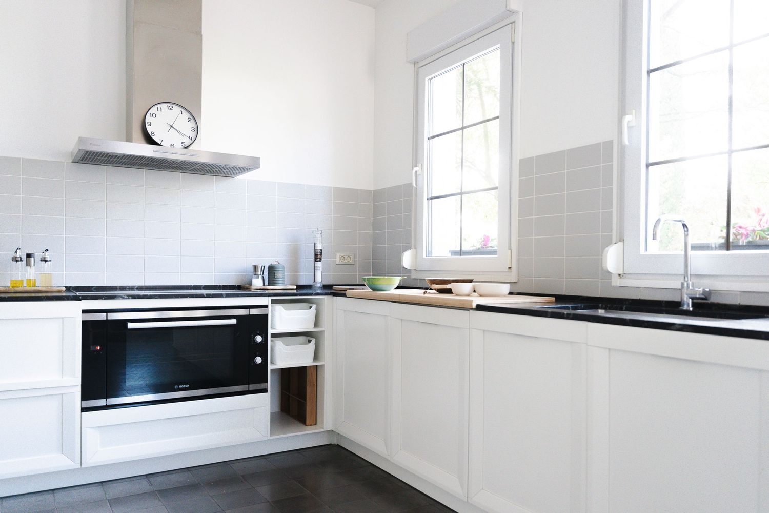 Modern white L-shaped kitchen with black countertops and stainless steel appliances features natural light from tall windows.