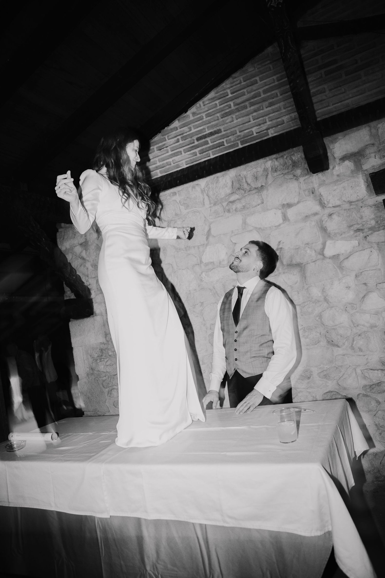 Black and white photo of couple dancing playfully on table during wedding reception.