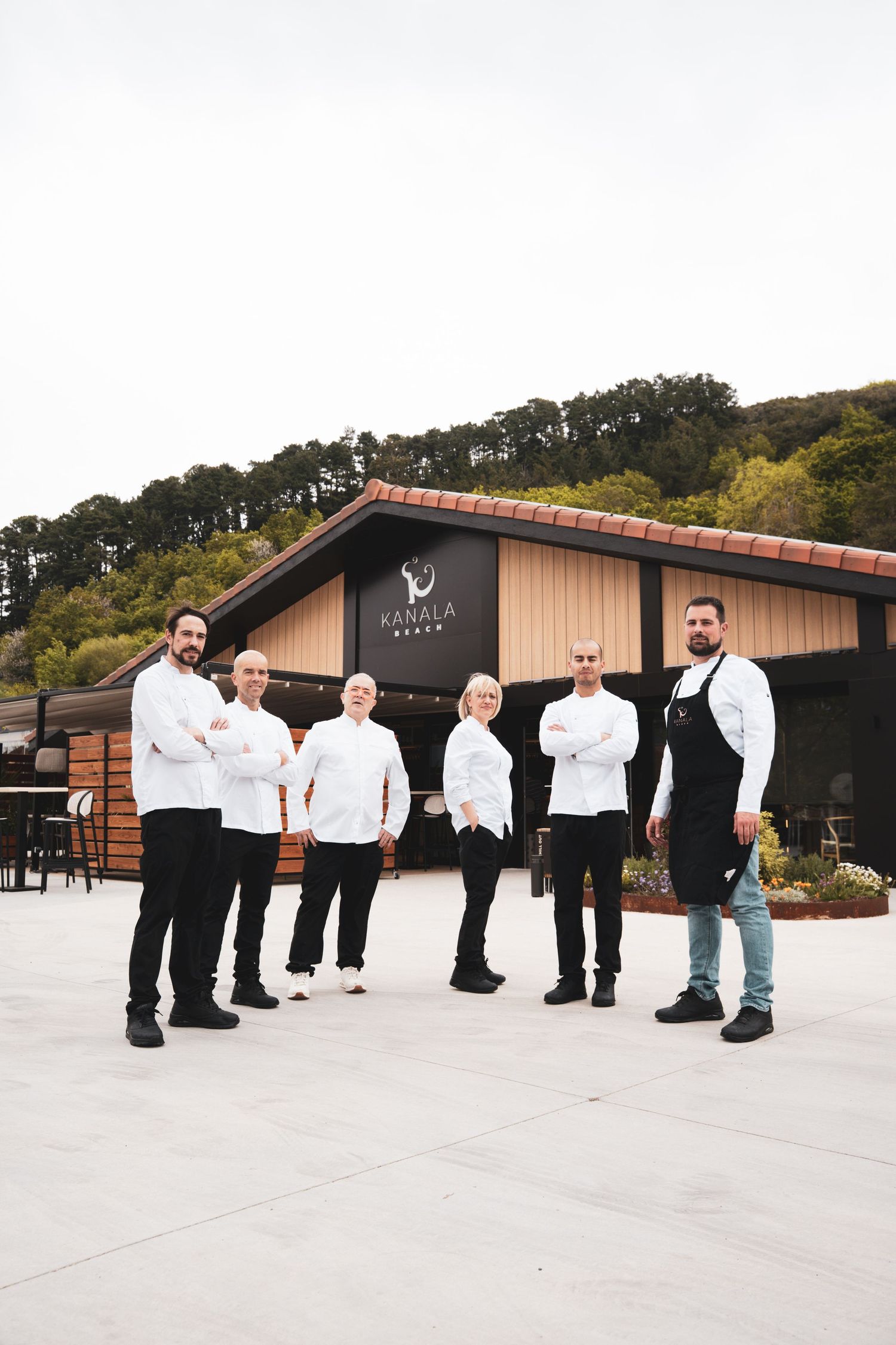Restaurant staff in white chef coats and black pants line up outside a modern building with wooden accents.