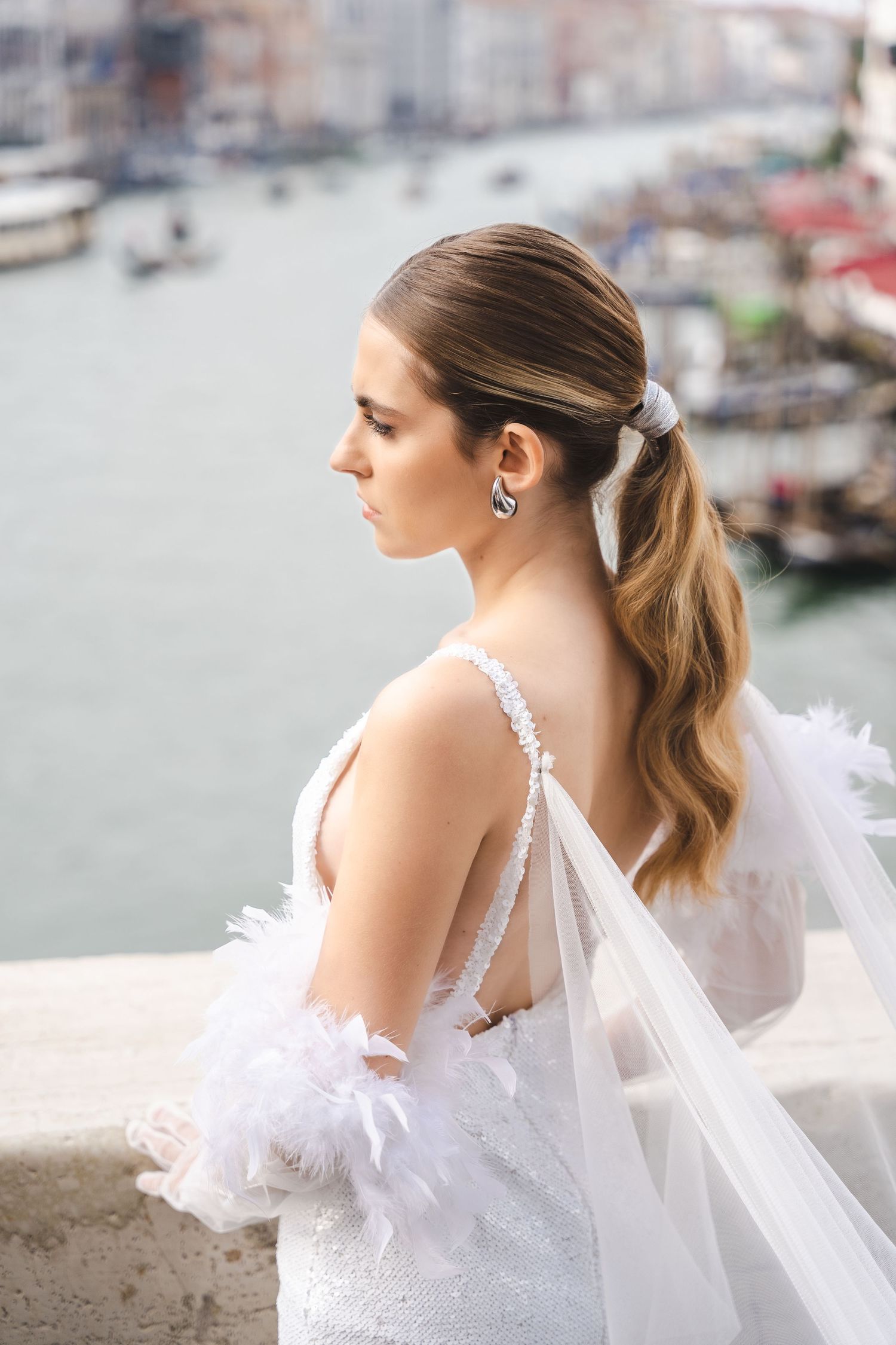 A bride in a white lace gown with flowing veil poses against a waterfront city backdrop.