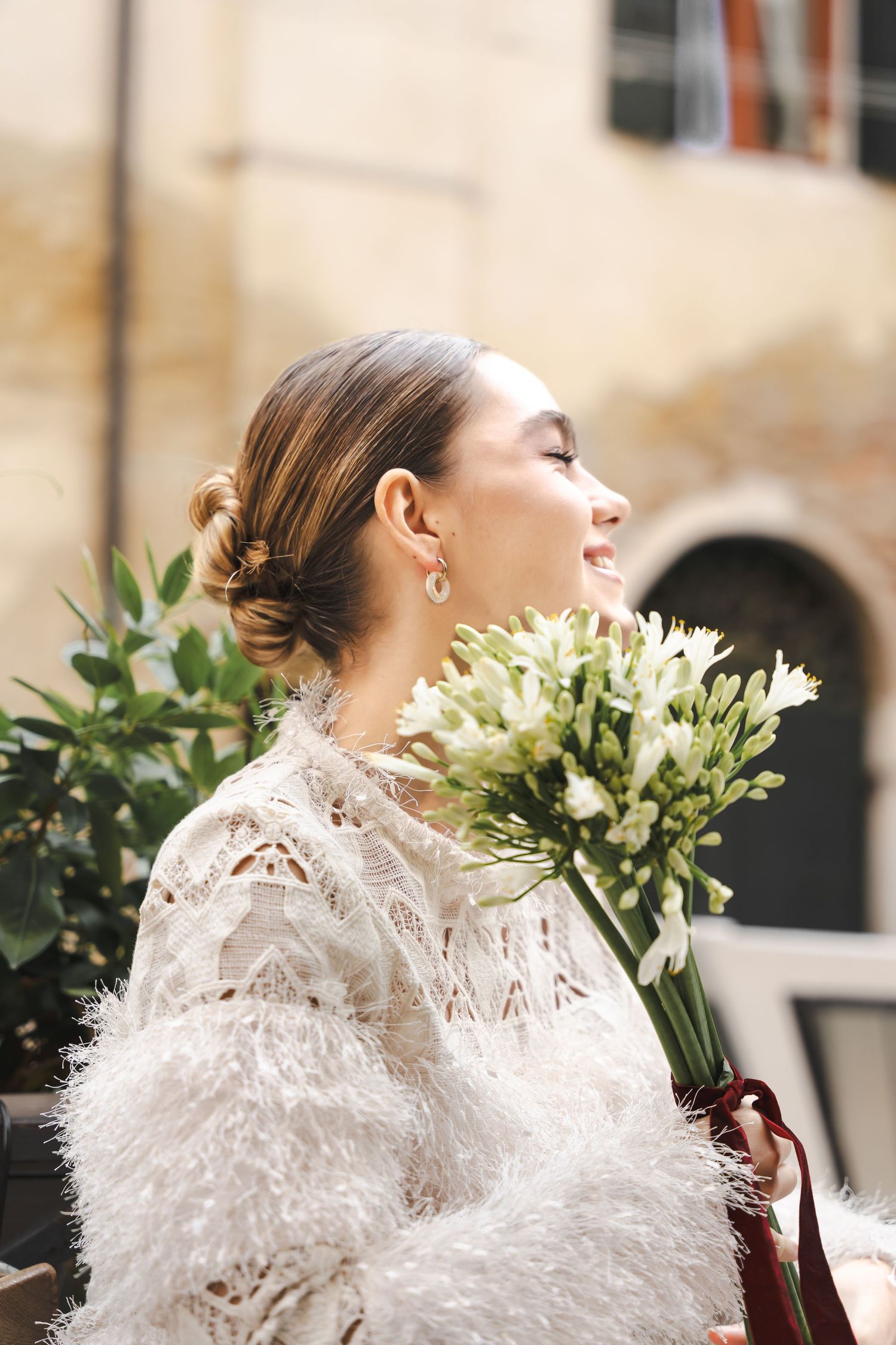 A bride in a delicate white lace and feather dress holds a bouquet of white lilies against a rustic exterior wall.