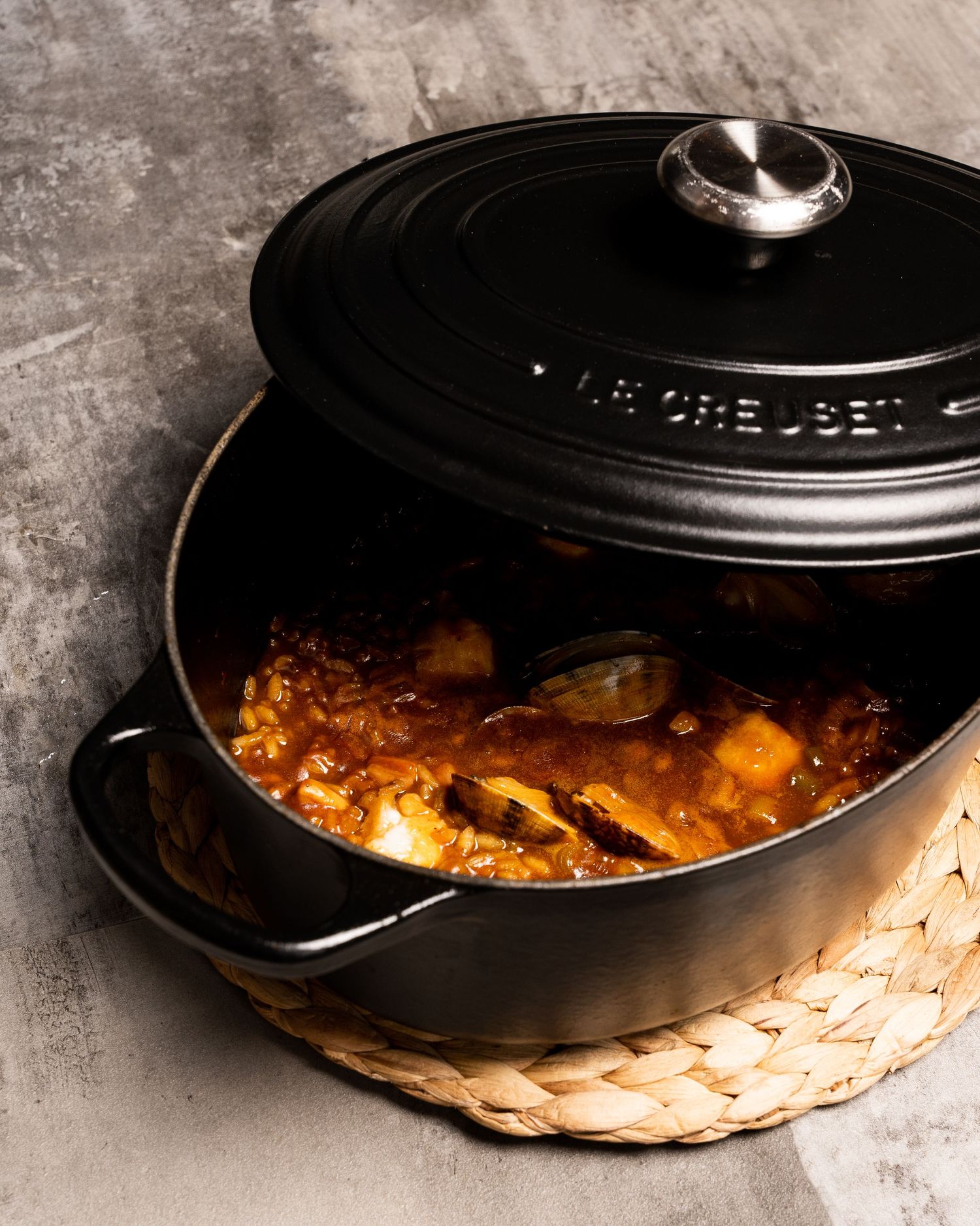A black Le Creuset Dutch oven filled with simmering stew sits on a woven trivet against a dark concrete surface.