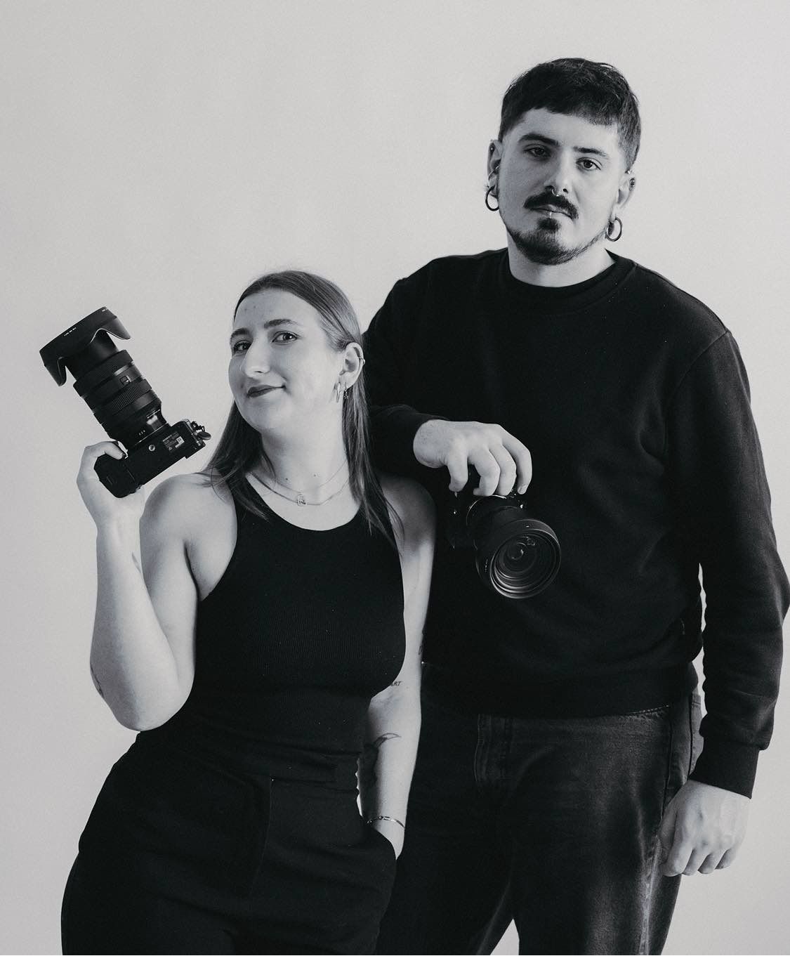 Photography duo in black outfits holding professional cameras against white background.