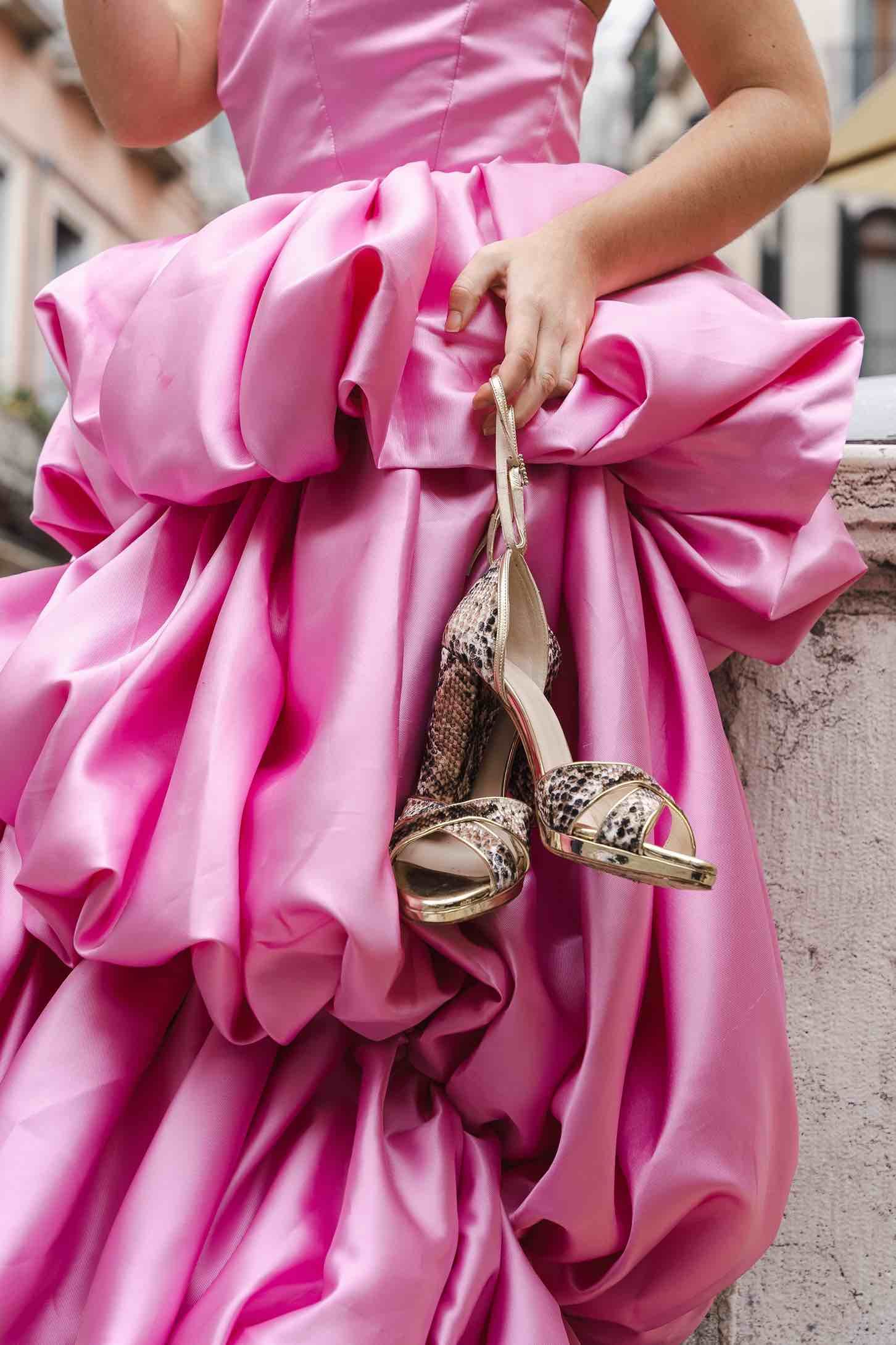 Pink satin ball gown with snakeskin heels held against architectural backdrop.