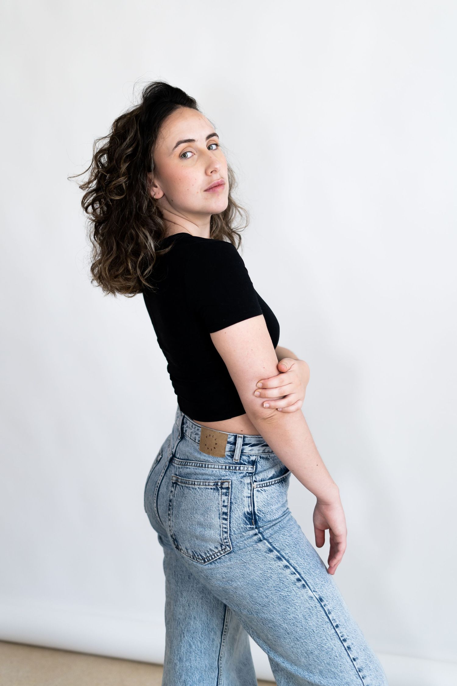 Fashion model in crop top and jeans poses with turned profile against white wall.