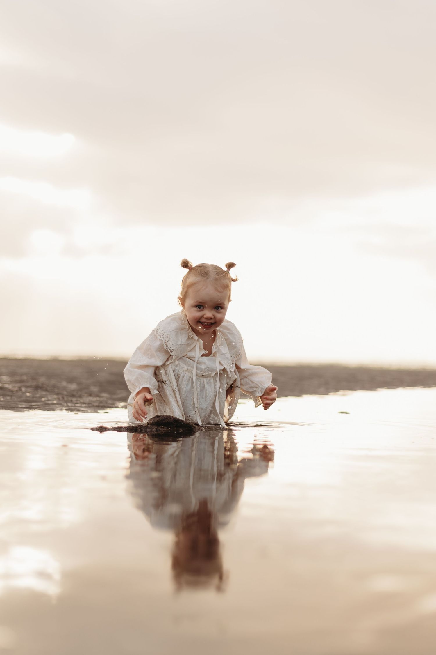 A small child wearing a white dress plays and sits in shallow reflective water on the beach at sunset.