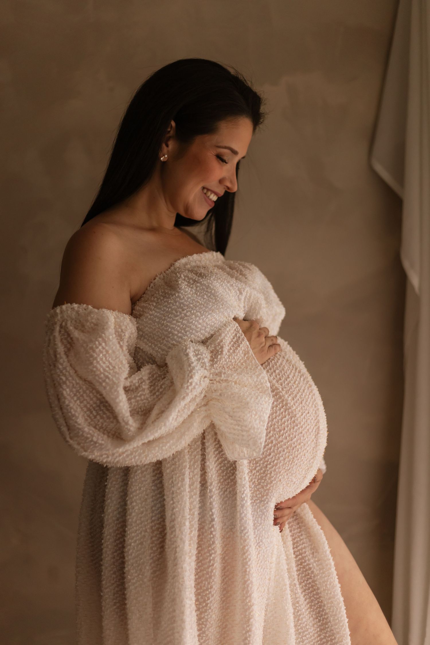 A person in a flowing off-shoulder white lace dress poses for an intimate maternity photo against a neutral backdrop.