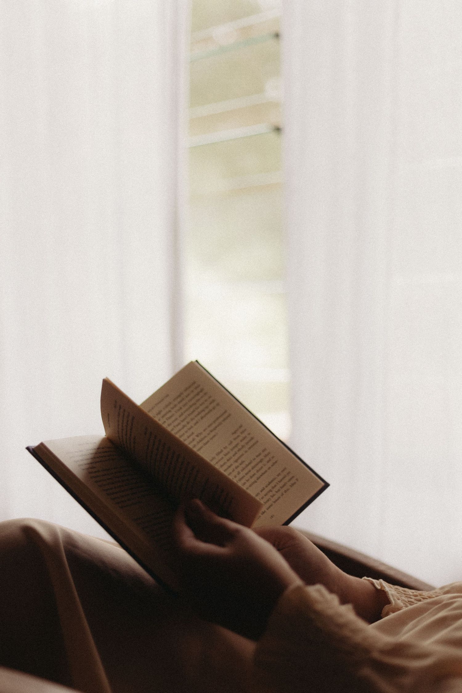 Hands hold an open book while relaxing in bed with soft natural lighting coming through a window.